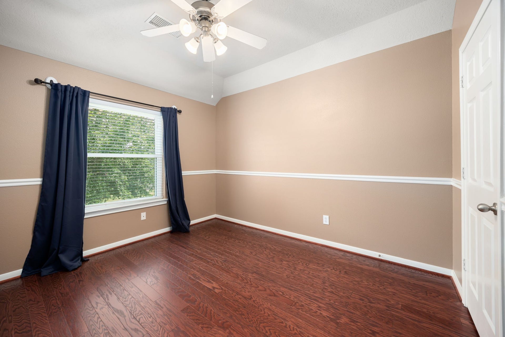 Additional upstairs secondary bedroom showcasing elegant chair rail molding and neutral wall tones. This inviting room is complete with premium wood-look flooring, a bright window overlooking green views, and a central ceiling fan.