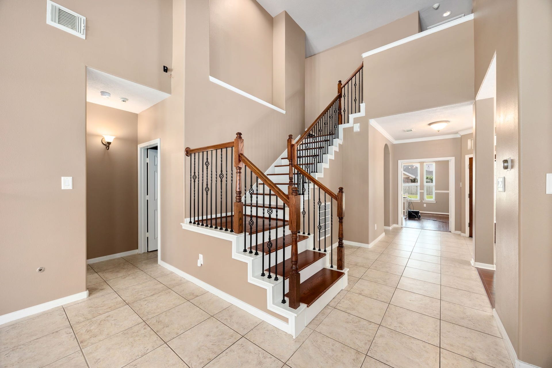 A striking central staircase serves as the focal point of the home, showcasing elegant wrought-iron spindles and rich wood treads. The soaring double-height ceilings and open-concept design create a dramatic, airy feel throughout the entryway.