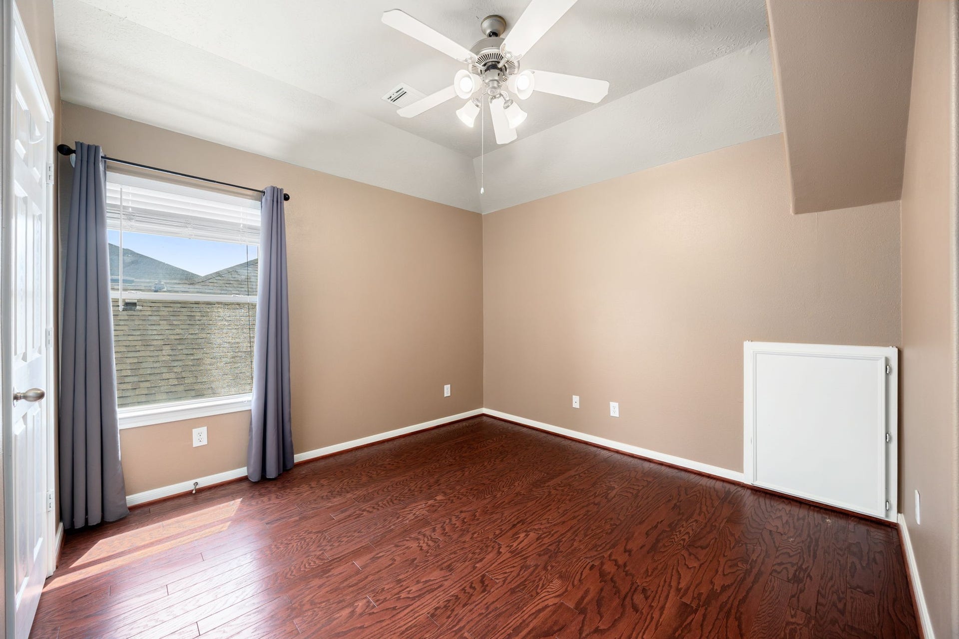 Charmingly appointed guest or children's bedroom featuring warm plank flooring and unique sloped ceiling lines. A large window fills the space with natural light, while a classic white ceiling fan ensures year-round comfort.