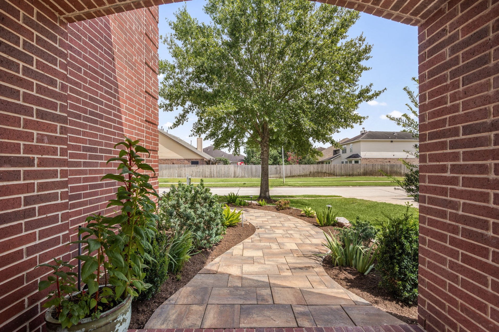 View from the covered entry revealing a beautifully winding paver walkway. The path is framed by professionally designed landscaping, lush garden beds, and a mature shade tree that enhances the front yard.