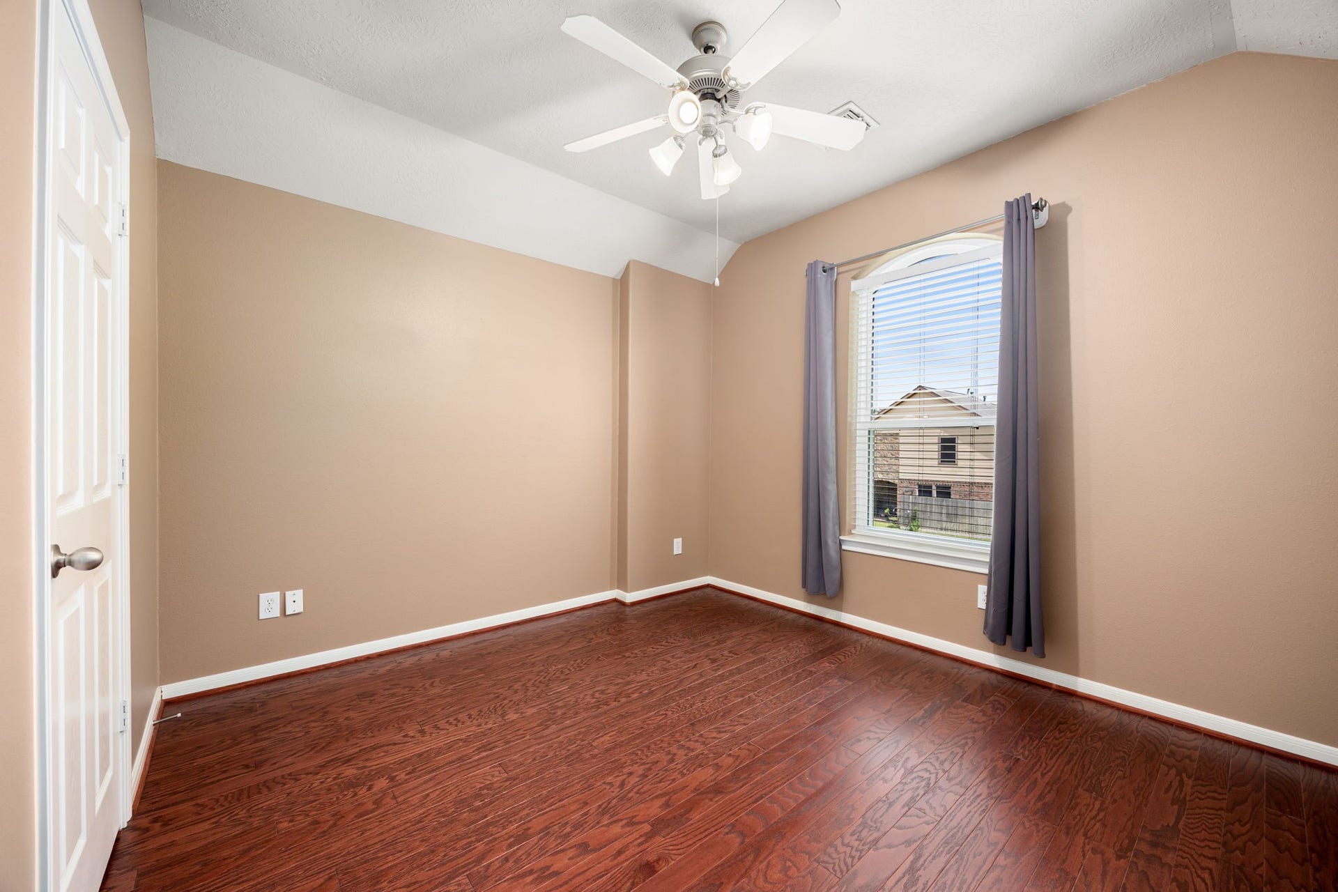 Generously sized secondary bedroom featuring rich wood-look flooring and a unique sloped ceiling accent. The room comes equipped with a modern ceiling fan, media-ready wall outlets, and a large arched window that lets in ample natural light.