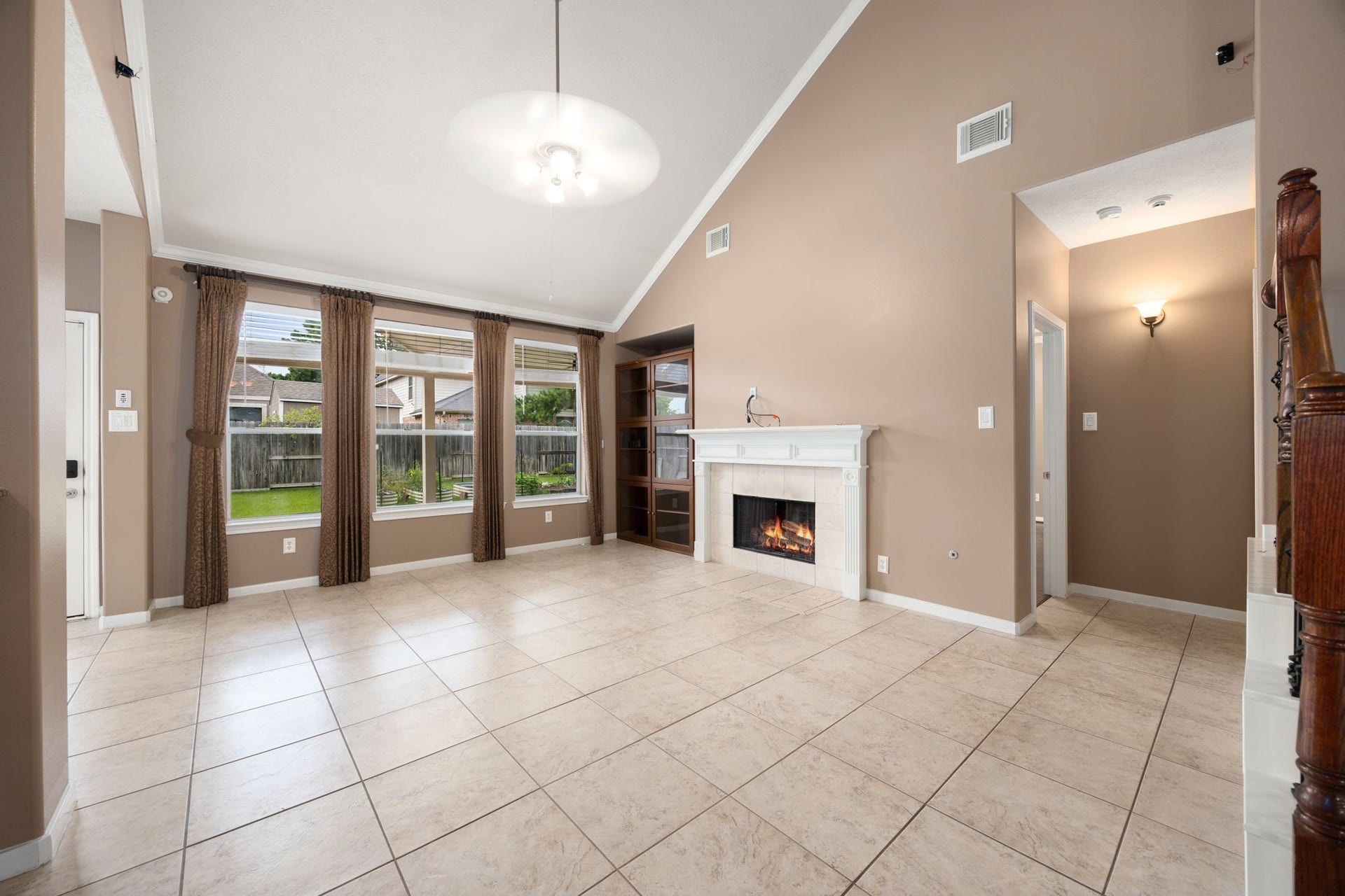Impressive family room boasting dramatic vaulted ceilings, low-maintenance tile flooring, and a bank of large windows looking out to the backyard. A cozy fireplace with a classic white mantel serves as the perfect centerpiece for gathering.