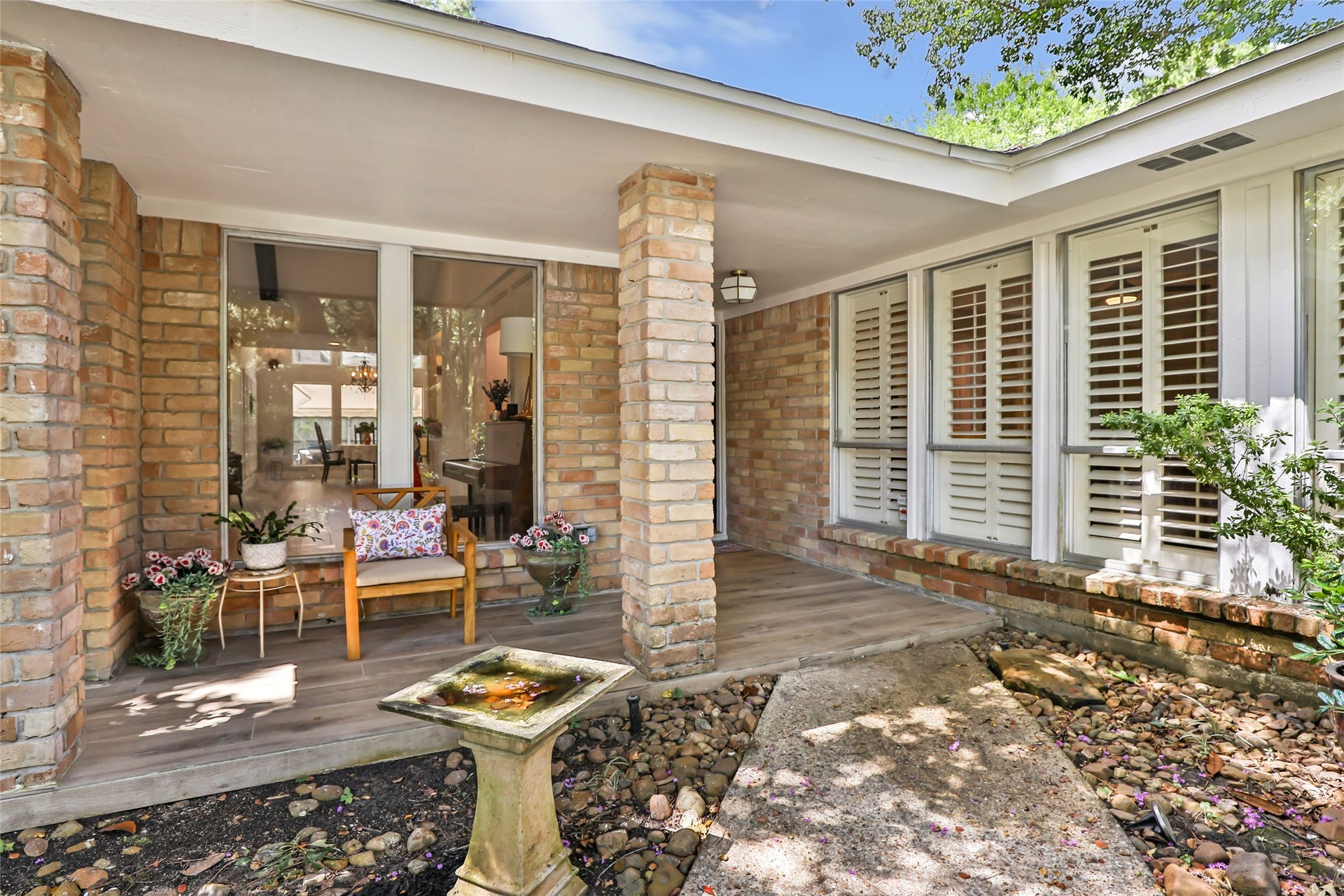 3714 Swift Creek Drive, Kingwood, TX 77339 Charming front porch with elegant brick columns, large windows with plantation shutters, and a cozy sitting area surrounded by lush greenery and a decorative birdbath.