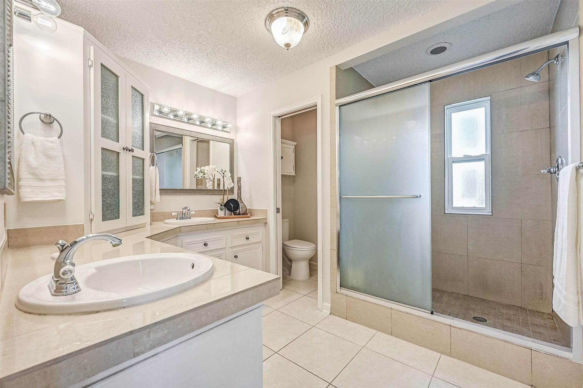 The primary bathroom features separate vanities with ample counter space, white cabinetry & a spacious tiled shower with a frosted glass enclosure. A window brings natural light into the shower, while a separate toilet area and additional storage cabinetry complete the space.