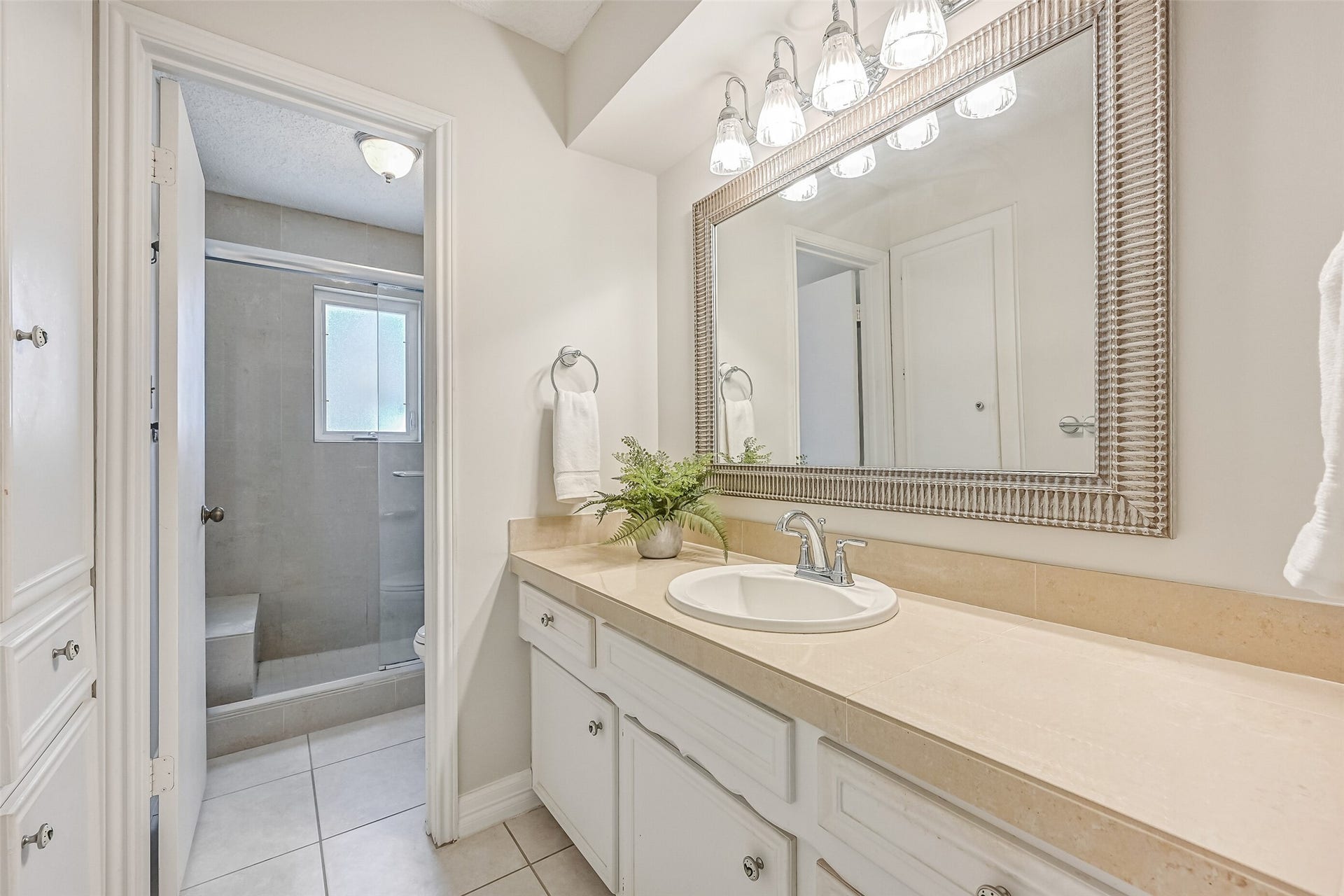 The secondary bathroom features a spacious vanity with light-colored countertop, white cabinetry, decorative framed mirror & updated light fixture. A separate bathing area includes a tiled tub/shower combination with glass enclosure and a window for natural light.