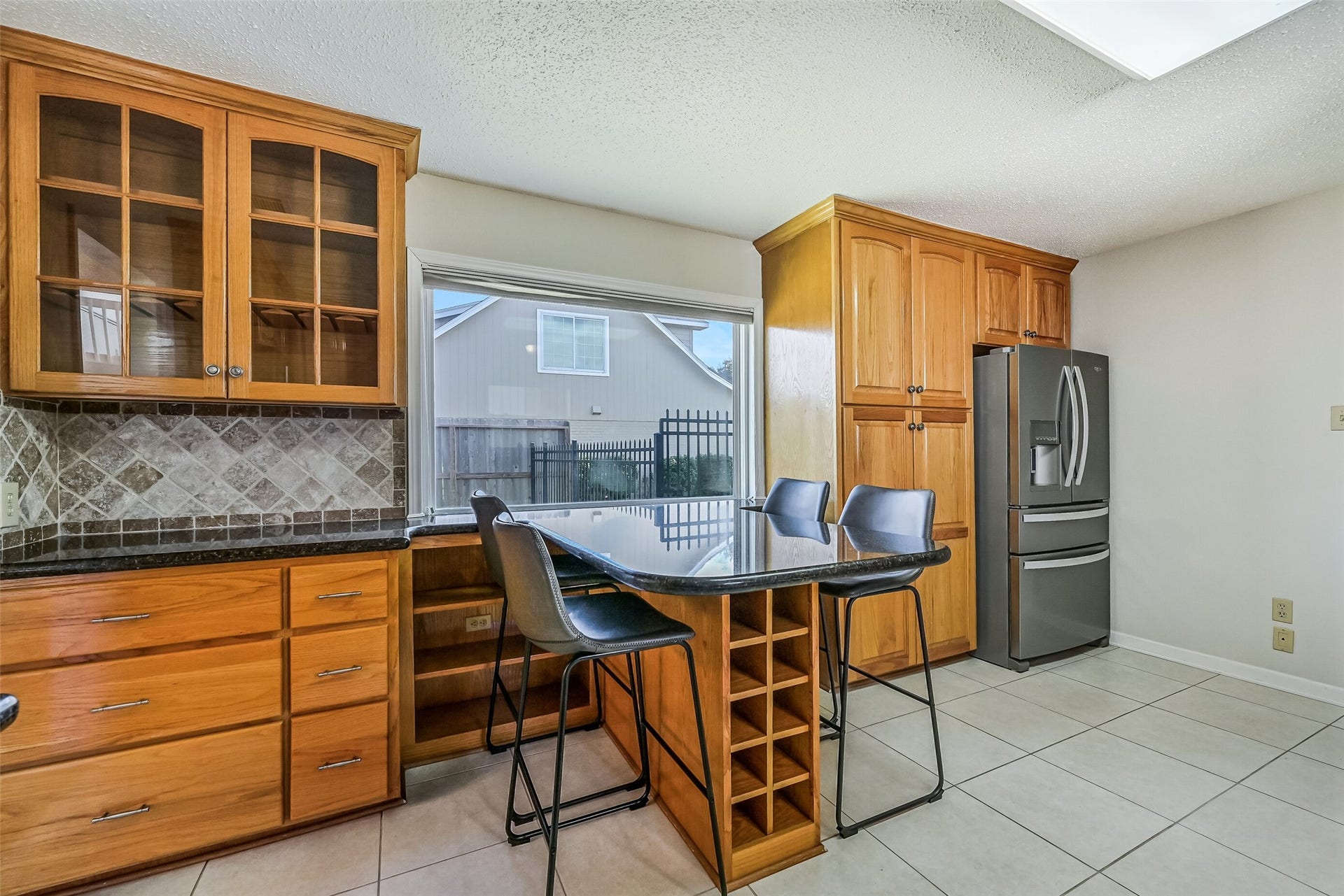 A granite-topped breakfast bar provides casual seating for four beside a large window overlooking the backyard. Built-in wine bottle storage, glass-front display cabinetry & additional shelving add character and functionality, while tall pantry-style cabinets offer plenty of storage space.