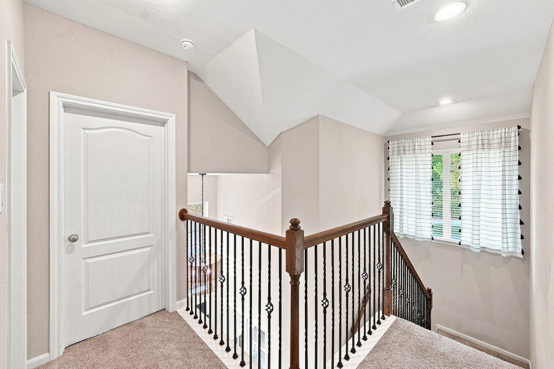 Bright and inviting upper hallway featuring a stylish staircase with wrought iron balusters, soft carpet underfoot, and a window with modern curtains providing natural light.