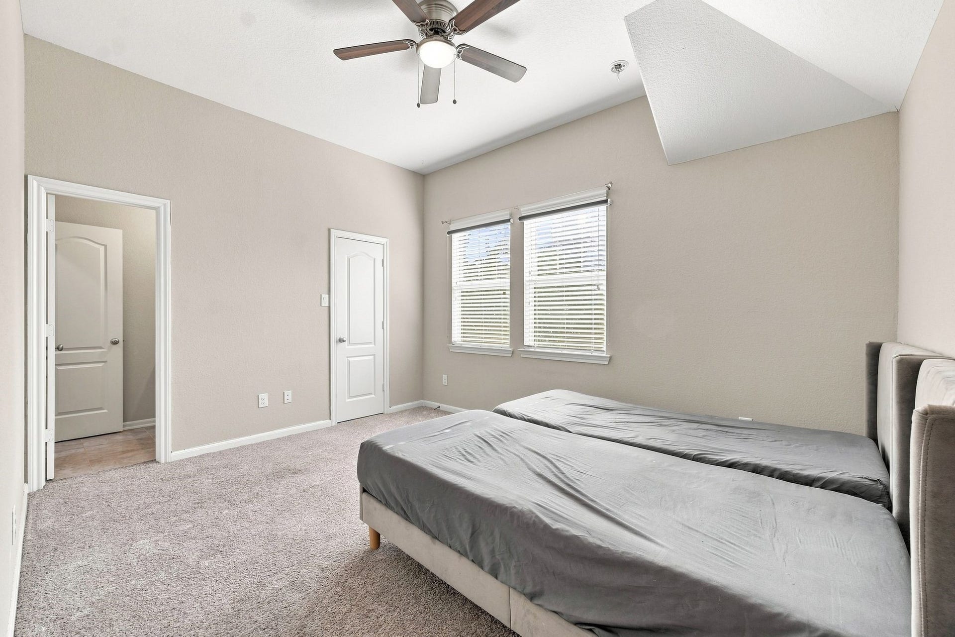 Spacious bedroom with soft carpet flooring, ample natural light from two large windows, and a modern ceiling fan for comfort.