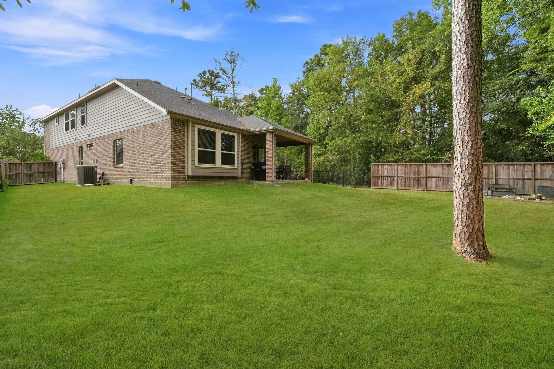 Spacious backyard featuring lush green lawn, a charming brick exterior, and a covered patio perfect for outdoor gatherings, all set against a backdrop of mature trees for added privacy.