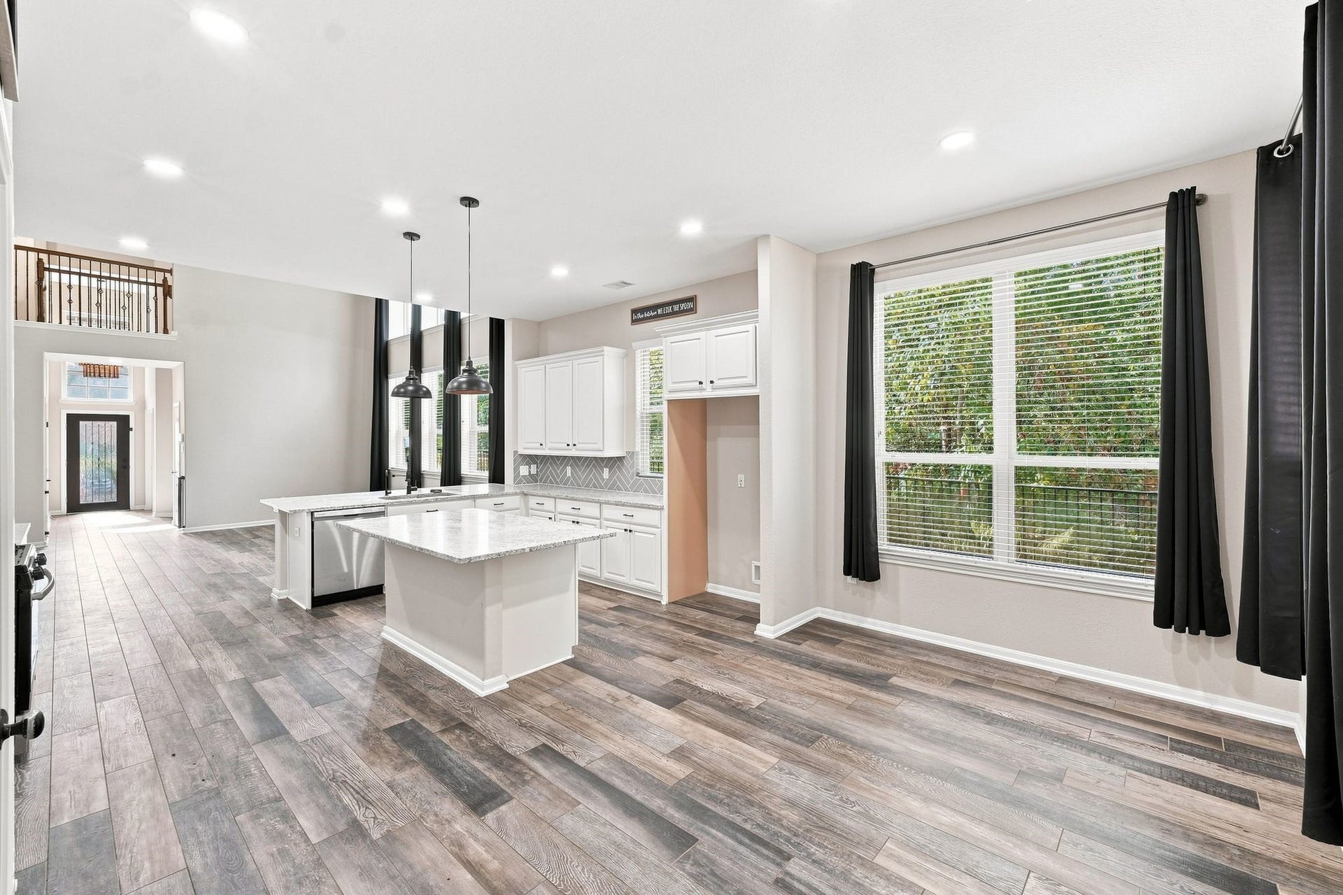 Spacious open-concept kitchen with modern white cabinetry, elegant pendant lighting, and a large island. Expansive windows invite abundant natural light, accentuating the sleek wood flooring and offering serene views of the greenery outside.