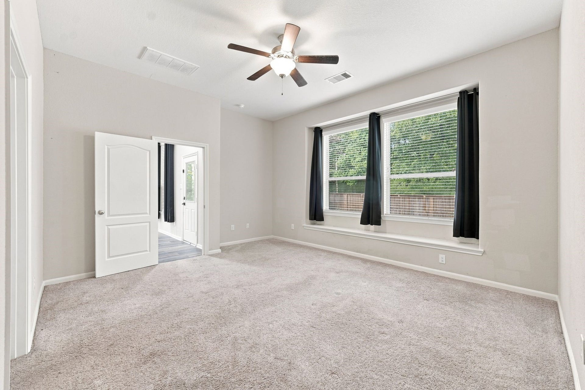 Spacious bedroom featuring plush carpeting, large windows with elegant drapes, and a ceiling fan for comfort.