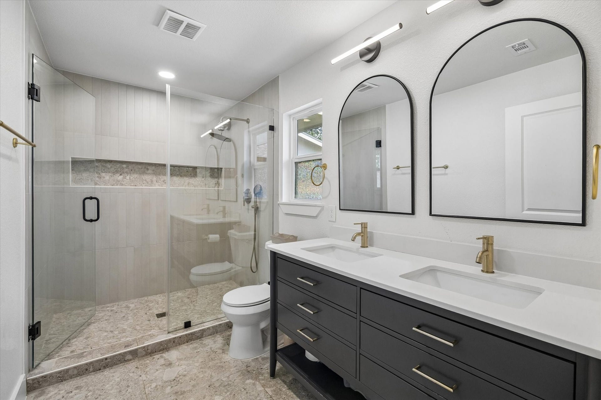 Clean lines give the primary bath a polished, modern feel, pairing a double vanity with warm brass accents and arched mirrors. An oversized glass enclosed shower features floor to ceiling tile, a built in niche, and handheld shower fixture.