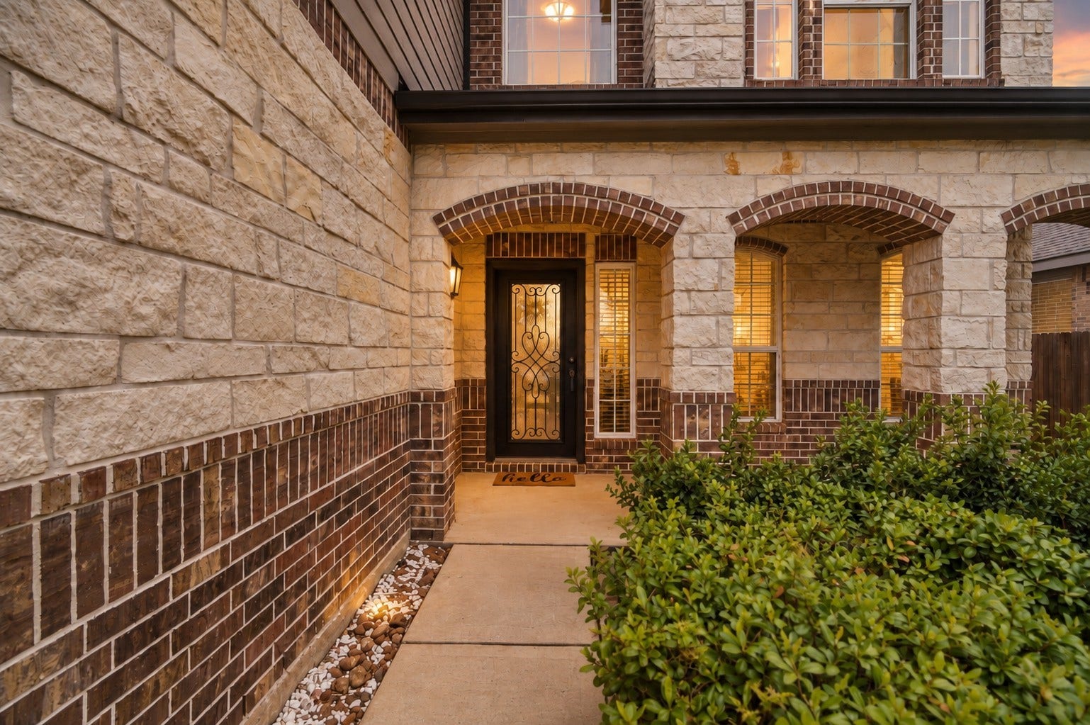 24115 Lake Molveno Drive, Katy, TX 77493 This inviting entrance features a grand covered porch crafted with textured stone masonry and rich brick accents. The design showcases elegant brick arches that frame tall sidelight windows, allowing warm indoor lighting to flow onto the concrete walkway.