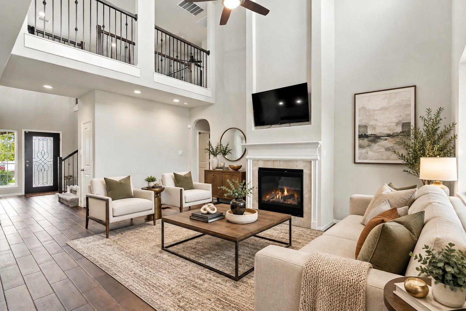 24115 Lake Molveno Drive, Katy, TX 77493 Virtually Staged: This dramatic, two-story living room boasts soaring ceilings and an upper-level mezzanine balcony featuring wrought-iron railing. The room is centered around a tiled fireplace with a mantel and TV accent wall.