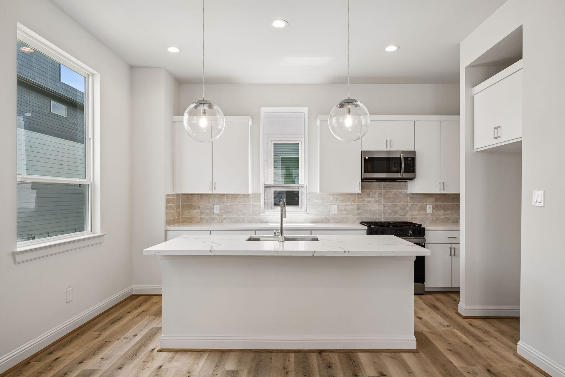 Elegant and modern, this kitchen boasts sleek white cabinetry, a spacious island with a stylish sink, and chic pendant lighting, all complemented by beautiful hardwood floors and ample natural light.