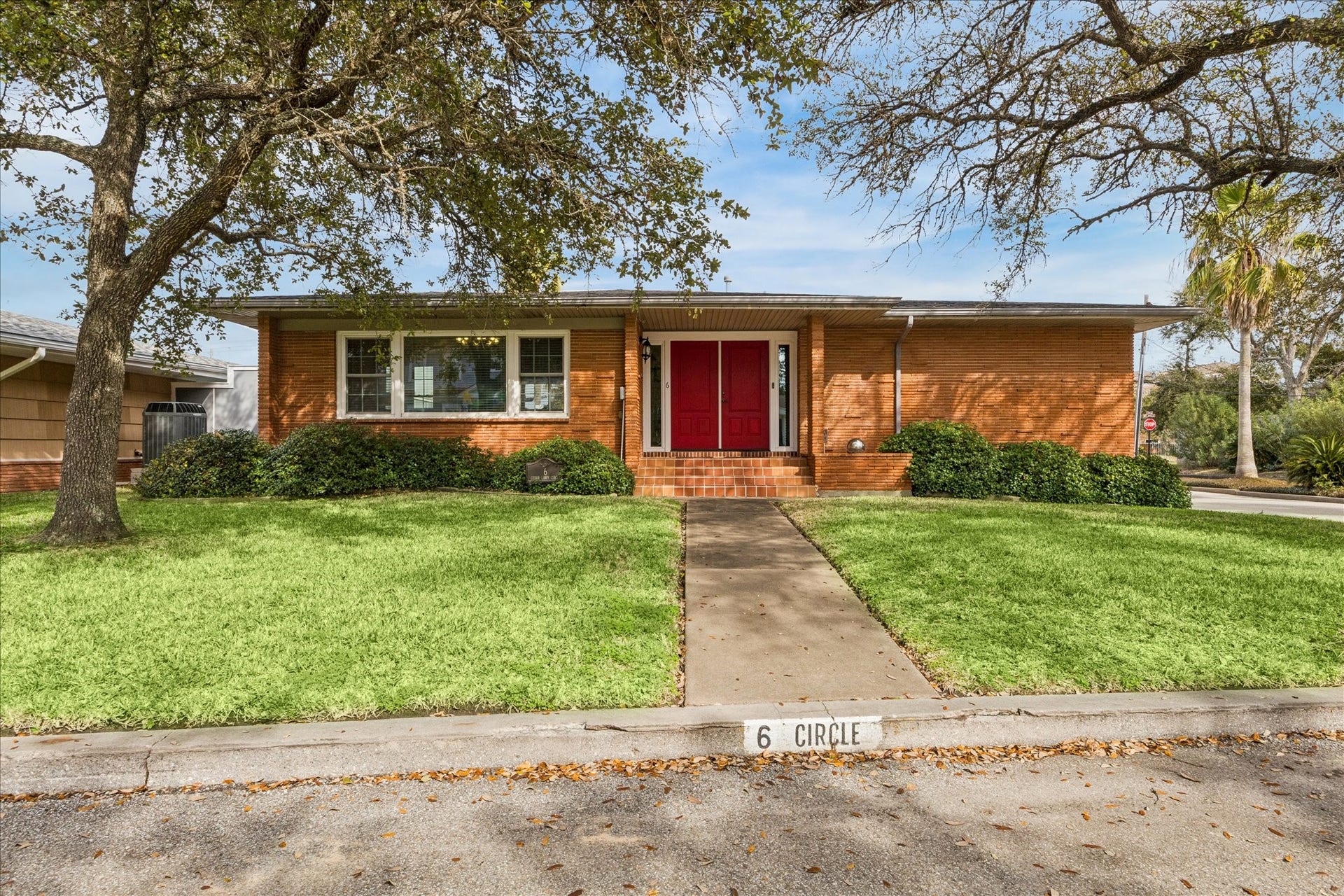 6 Cedar Lawn Circle, Galveston, TX 77551 Charming brick home with a welcoming red front door, set on a corner lot. Features a manicured lawn, mature trees, and a simple concrete pathway leading to the entrance.