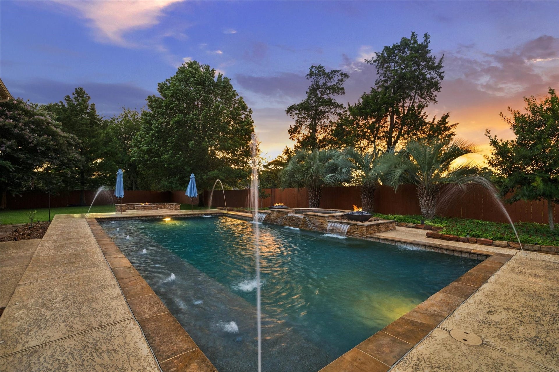 24935 Waterstone Estates Circle W, Tomball, TX 77375 A stunning poolside retreat surrounded by mature trees and lush landscaping, where dramatic water features and glowing fire accents create a serene and inviting atmosphere at sunset.
