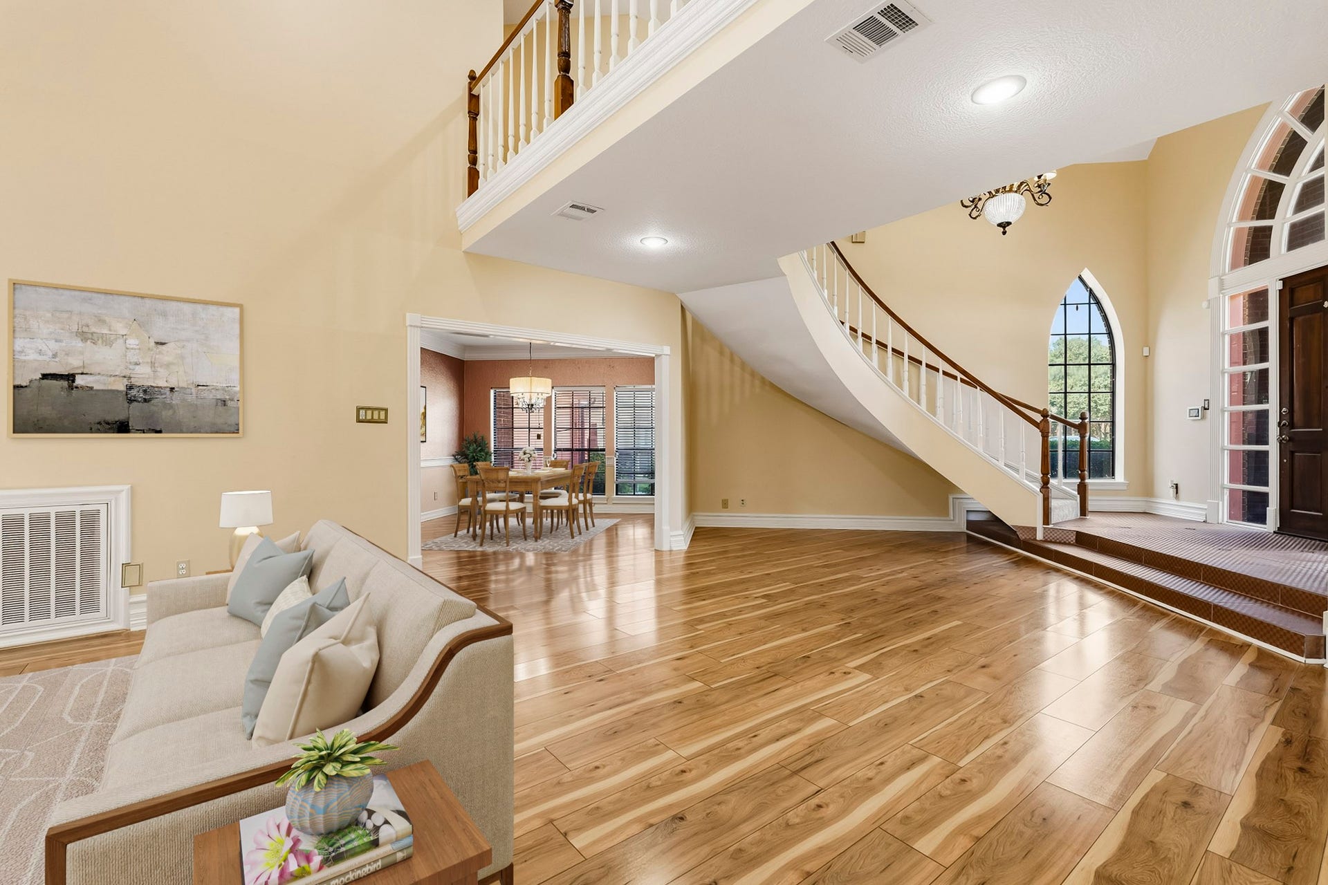 Virtually Staged: Open-concept living room featuring gleaming hardwood floors and high ceilings. Looking past the curved staircase, a wide cased opening connects seamlessly into the formal dining room.