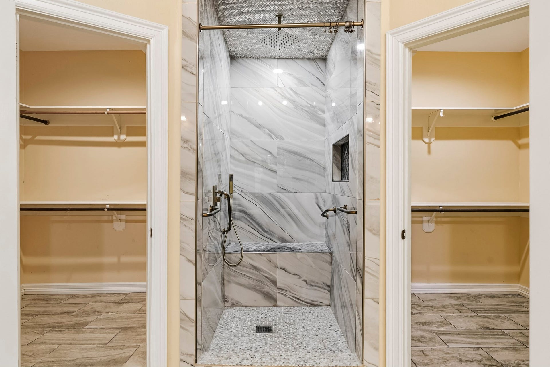 Highlights a walk-in shower with marble-look surround, rain showerhead, mosaic floor, and built-in bench seating, perfectly centered between two spacious walk-in closets.