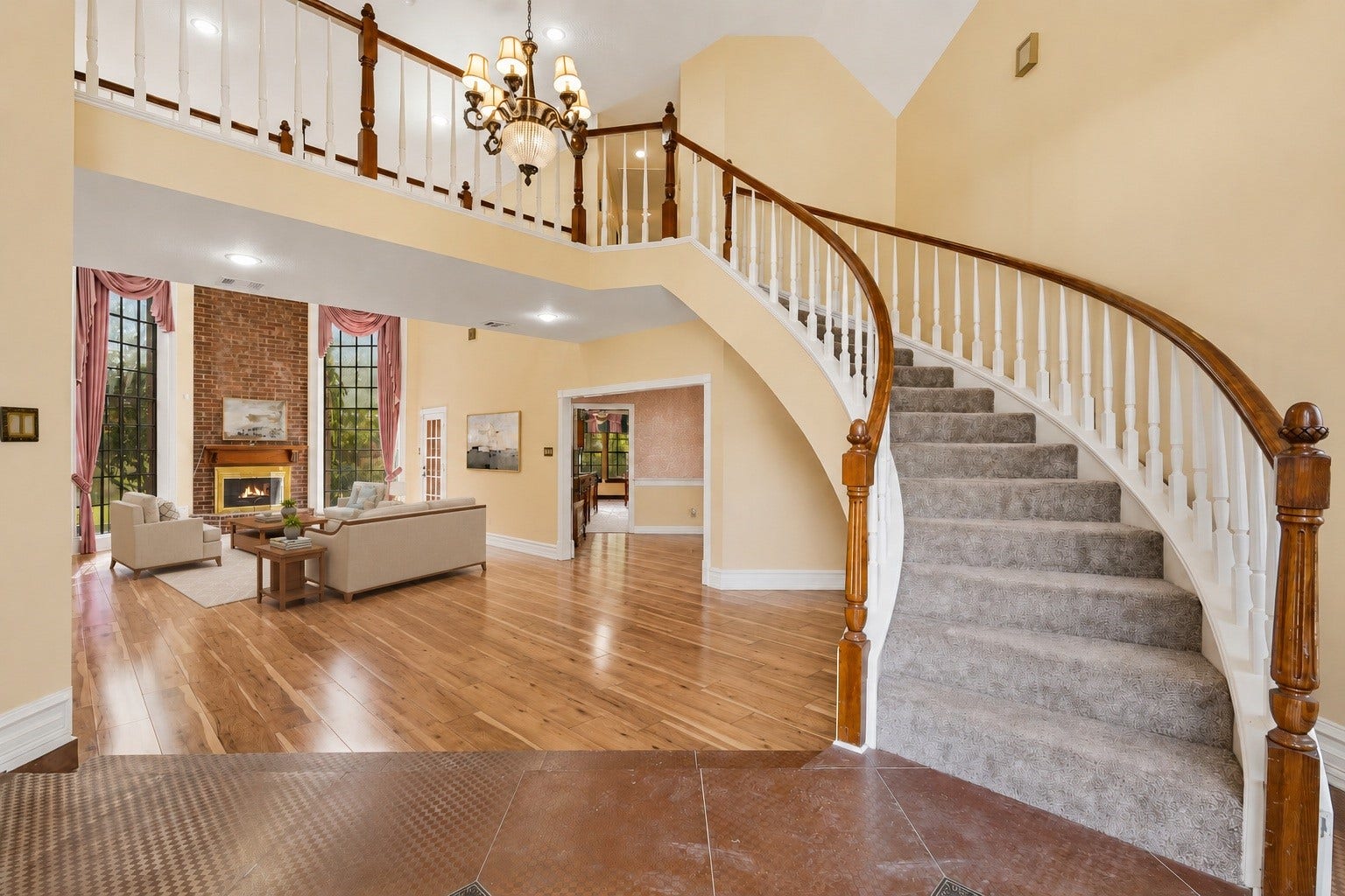 Virtually Staged: Welcome home to a grand foyer with a graceful curved staircase, gleaming hardwoods, and a sunlit living room anchored by a brick fireplace and soaring windows.