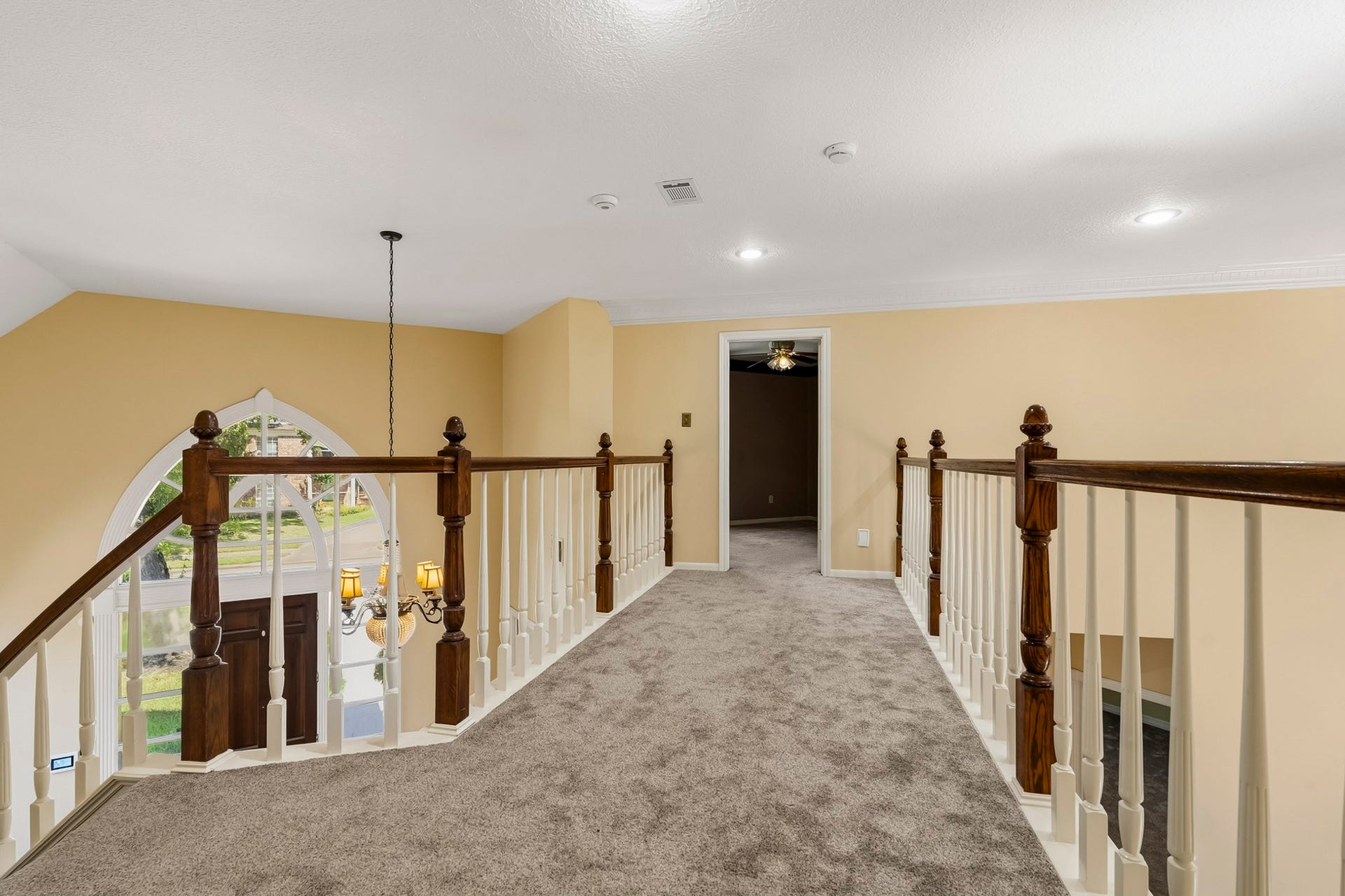 Impressive second-floor catwalk with carpeted flooring and wooden banisters overlooking the two-story entryway, filled with natural light from the grand arched window below.