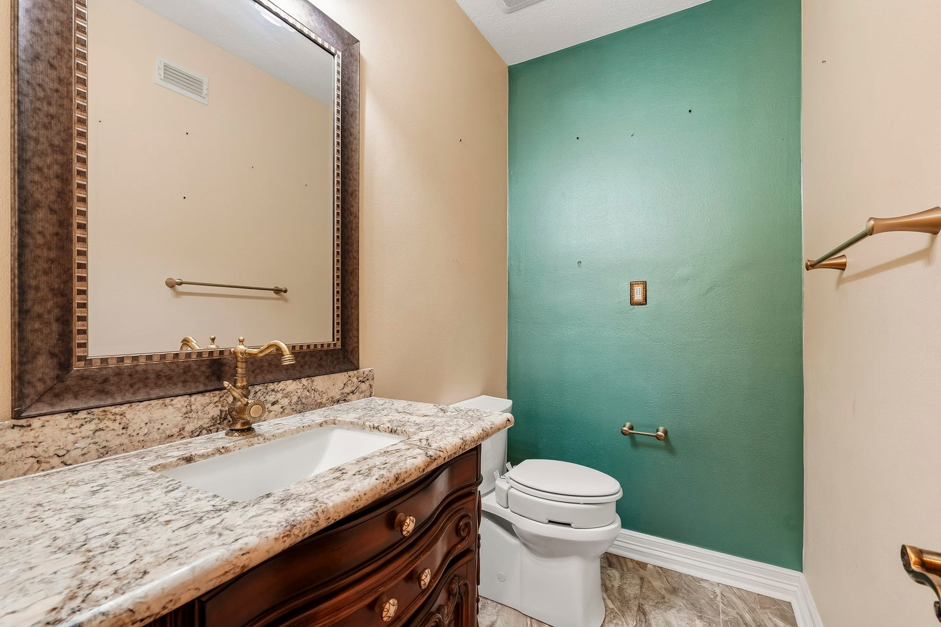 Elegant half bath equipped with a classic furniture-style vanity, granite countertop, ornate mirror, vintage-inspired brass hardware, and tile flooring for guests.