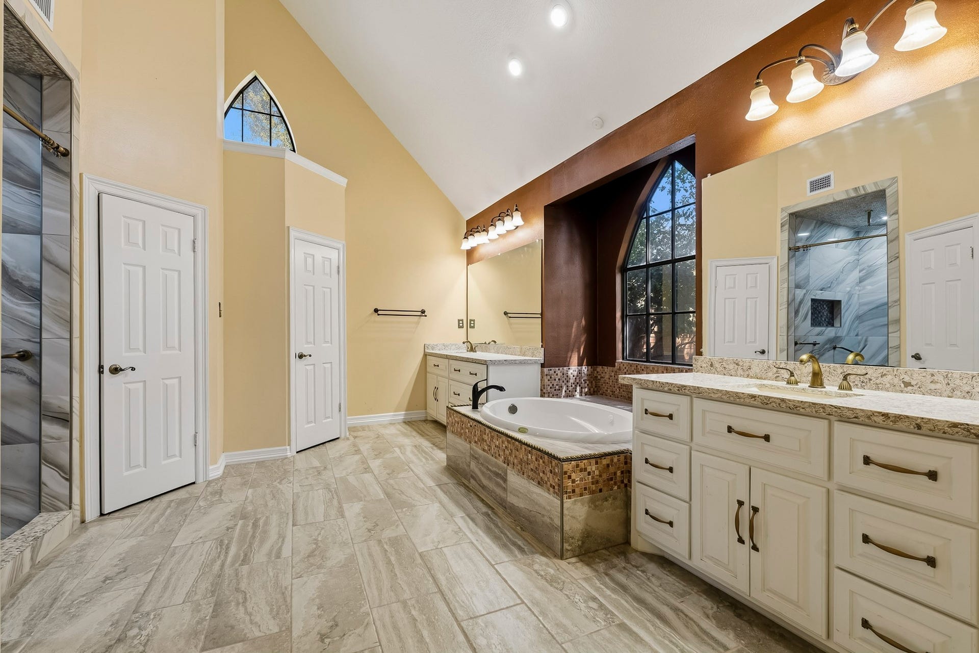 Luxurious primary bath featuring vaulted ceilings, dual vanities, and stone tile floors. Enjoys a soaking tub set beneath a dramatic arched window, a large walk-in shower, and a spacious walk-in closet.