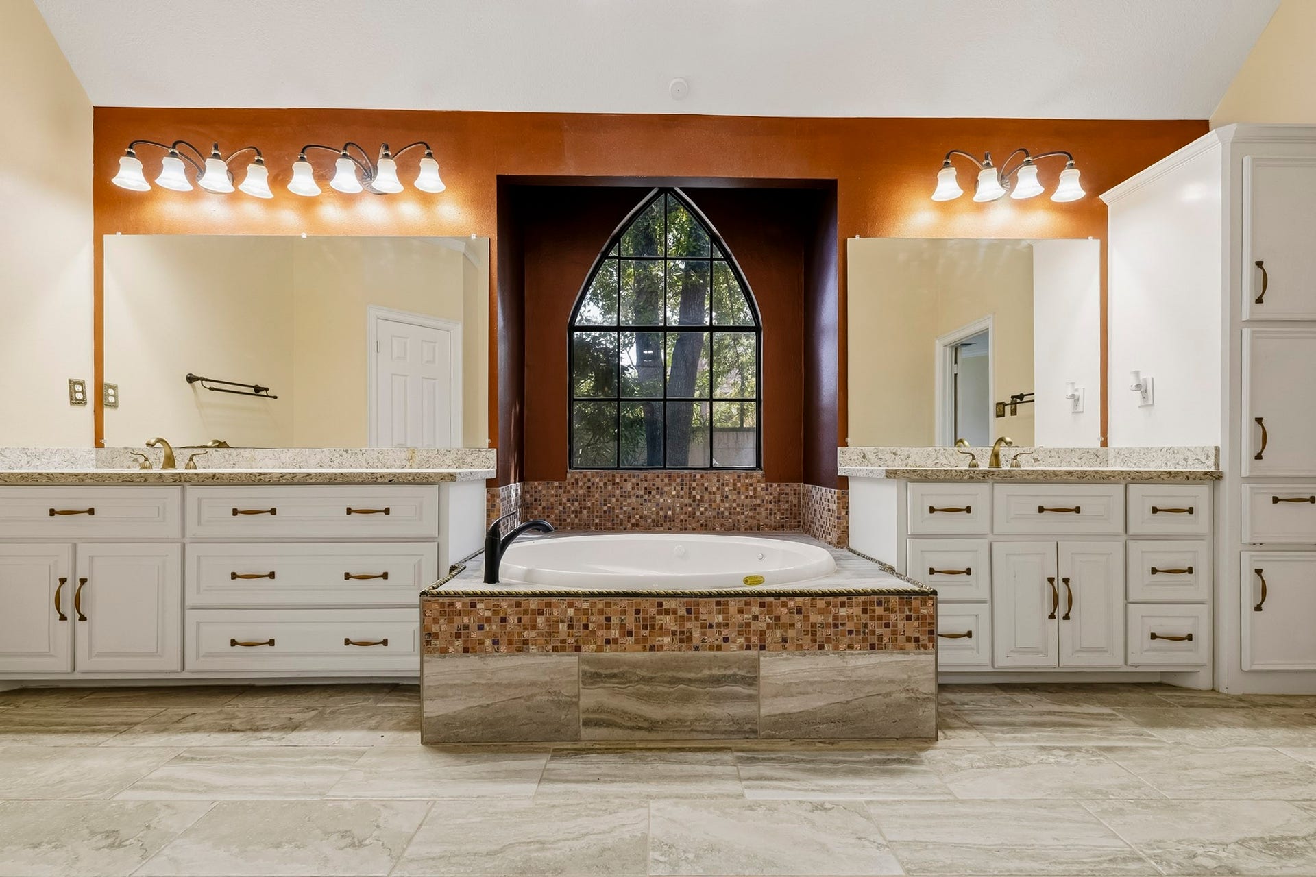 Stunning centered shot of the primary bath! Features split dual vanities, a central soaking tub under an elegant arched window.