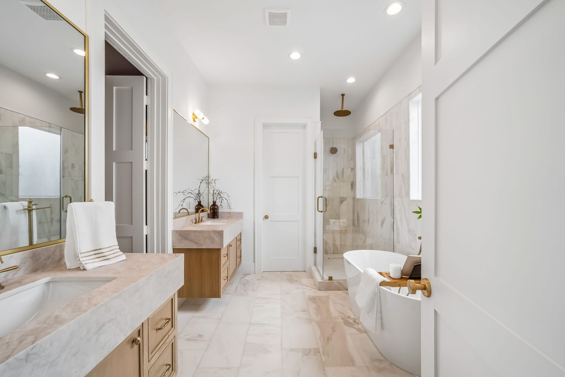 First-floor en-suite bathroom featuring a freestanding soaking tub, a glass-enclosed walk-in shower with elegant gold fixtures, dual vanities with leathered Taj Mahal quartzite countertops, floating white oak cabinetry, and timeless marble flooring, creating a refined and luxurious retreat.