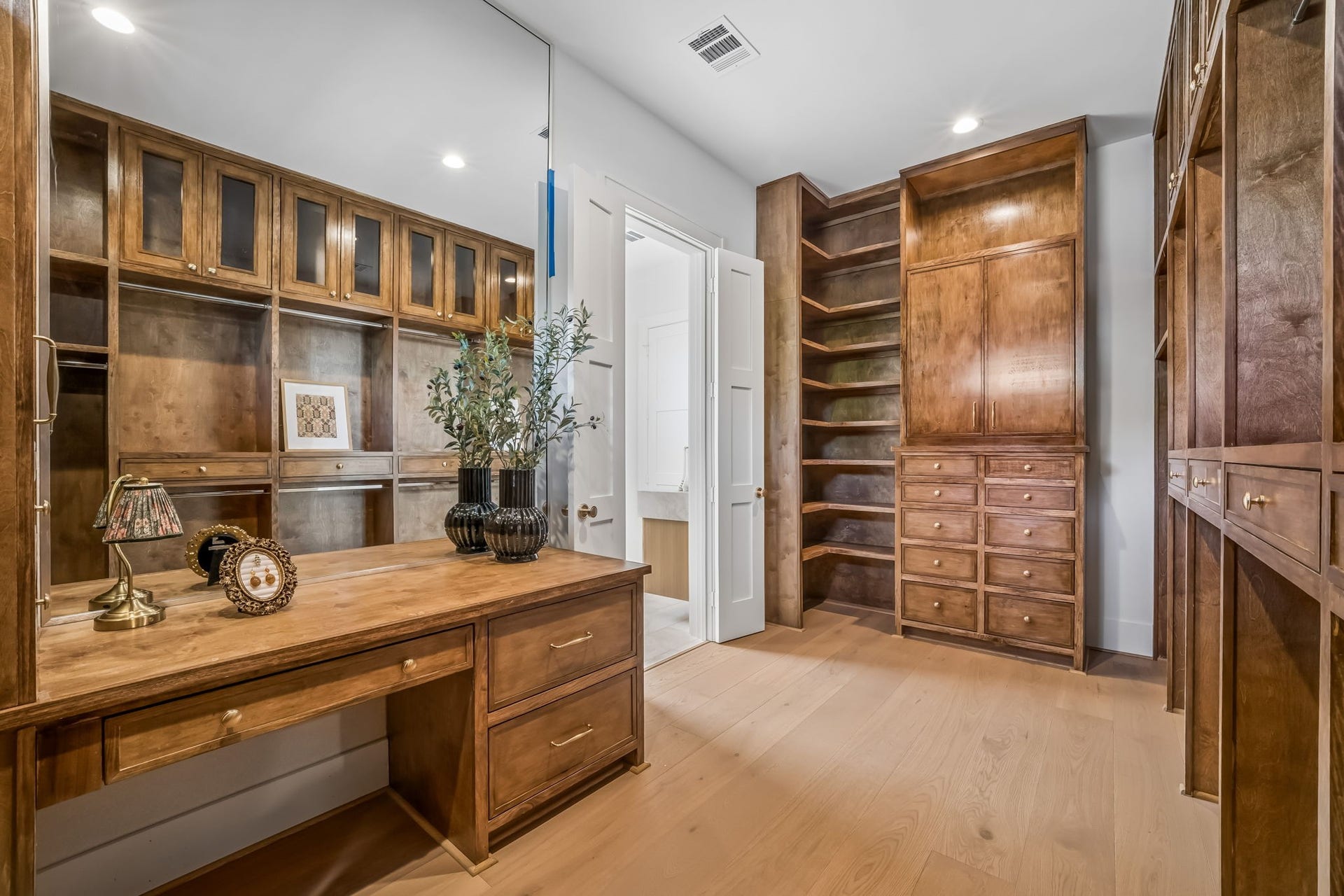 The custom closet of your dreams, designed with a dedicated vanity area, floor-to-ceiling storage, glass-enclosed cabinetry, and an oversized mirror.