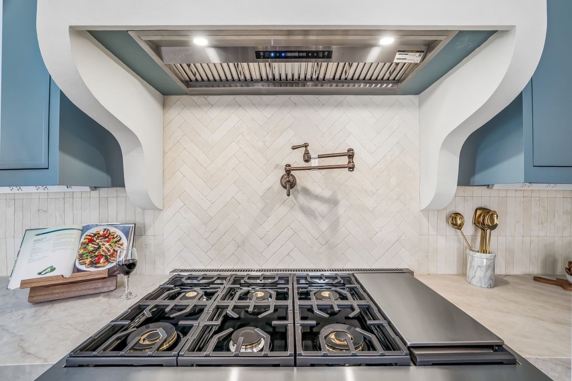 Custom chefs kitchen featuring a professional-grade gas range, stainless steel vent hood, elegant herringbone tile backsplash, and striking blue cabinetry with refined brass accents.
