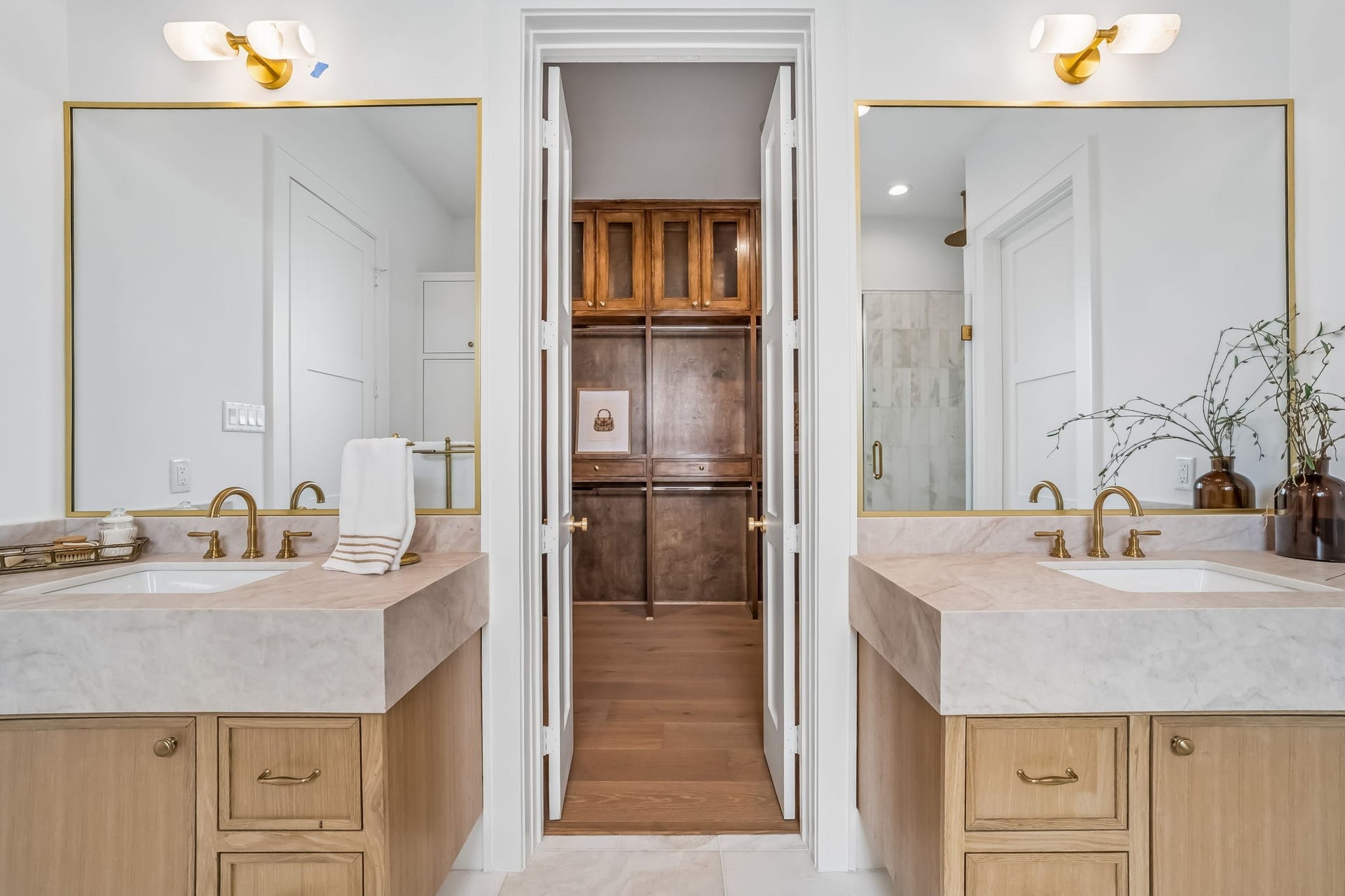 Elegant luxury primary bathroom showcasing dual vanities with marble countertops, custom wood cabinetry, gold fixtures, oversized mirrors, and refined finishes, with a private walk-in closet beyond.