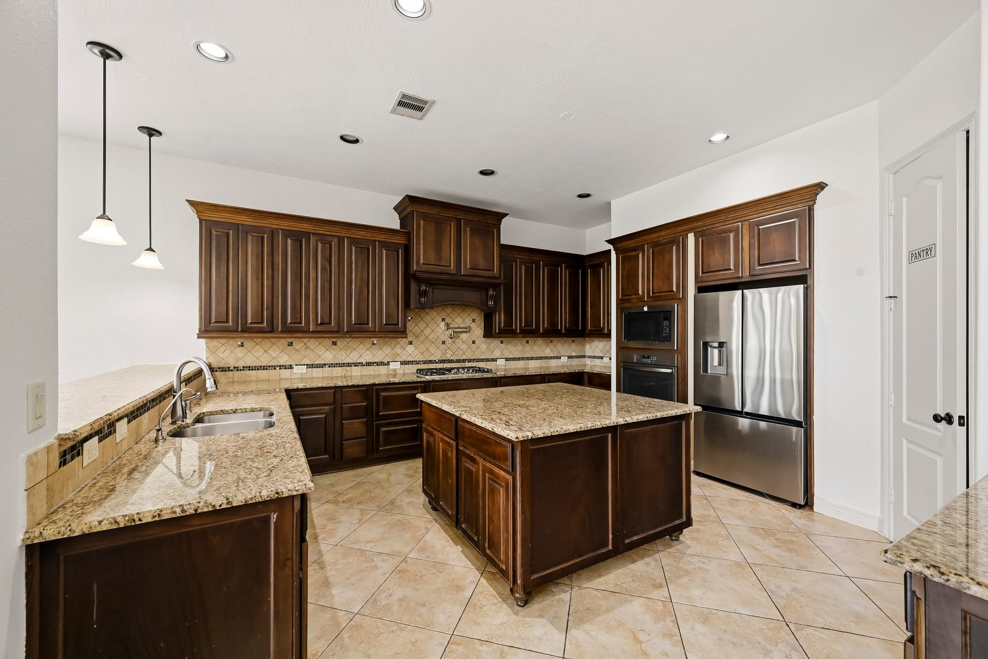27730 Dalton Bluff Court, Katy, TX 77494 This spacious kitchen features rich dark wood cabinetry, granite countertops, and a large center island that provides generous prep space and additional storage. Stainless steel appliances, including a French-door refrigerator, built-in microwave, and oven, complement the warm finishes. A decorative tile backsplash, double-basin sink, gas cooktop, and abundant cabinetry add functionality, while pendant and recessed lighting keep the space bright. A dedicated walk-in pantry provides additional storage.