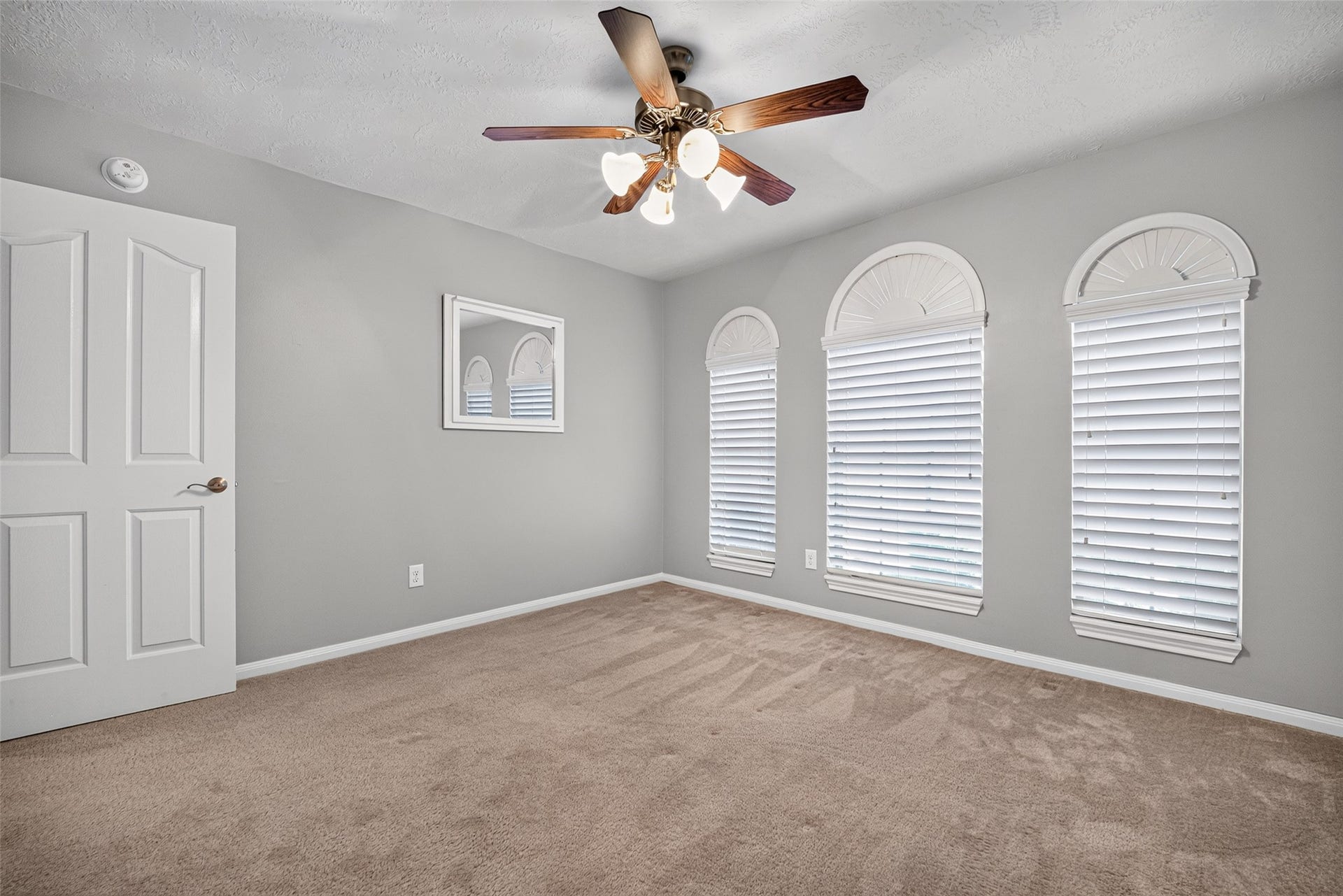 Secondary bedroom features three arched windows with white blinds, a ceiling fan, carpeted flooring, and a paneled entry door.
