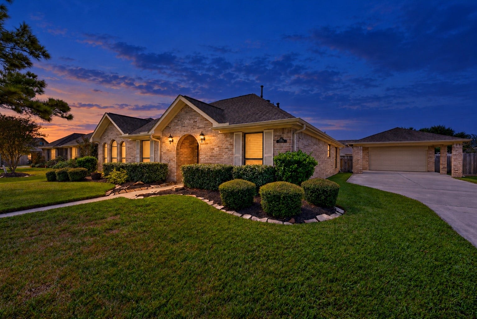Virtually enhanced twilight photo highlights the brick exterior, arched front entry, landscaped beds, and driveway leading to the detached garage.