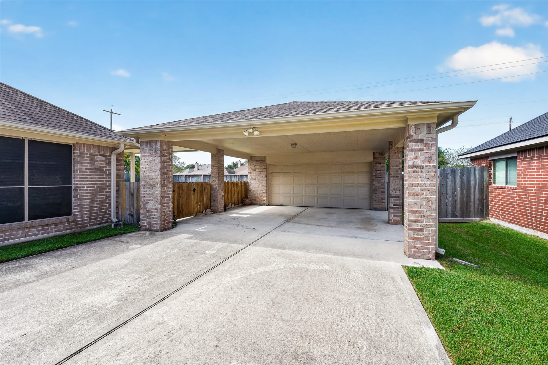 Concrete driveway leads beneath a broad covered parking area supported by brick columns, with a garage door at the rear.