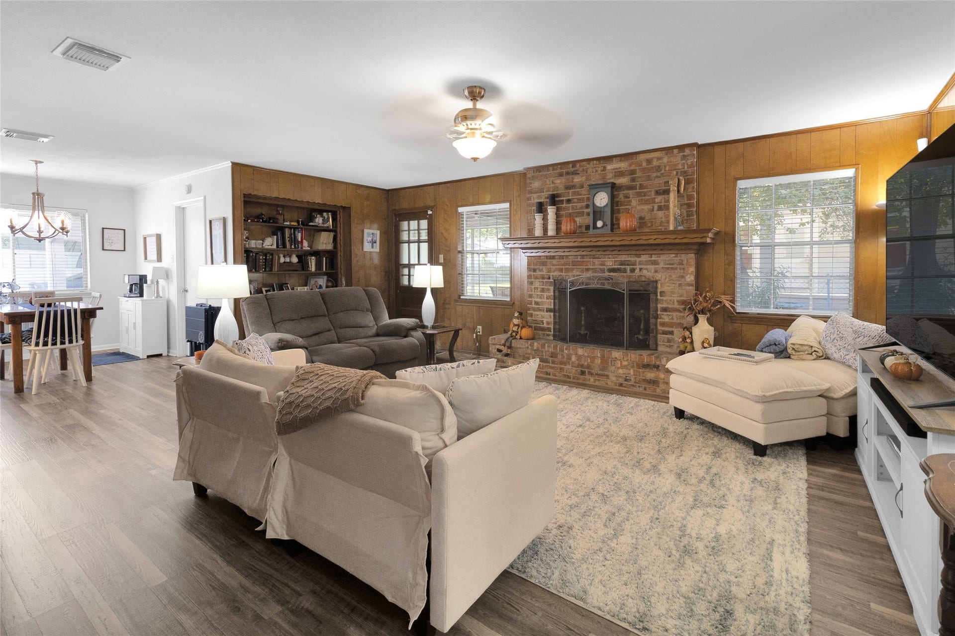 613 N Avenue F, Humble, TX 77338 Charming living room with a cozy brick fireplace, rich wood paneling, and ample natural light. The open layout seamlessly connects to the breakfast area, offering an inviting space for relaxation and entertaining.