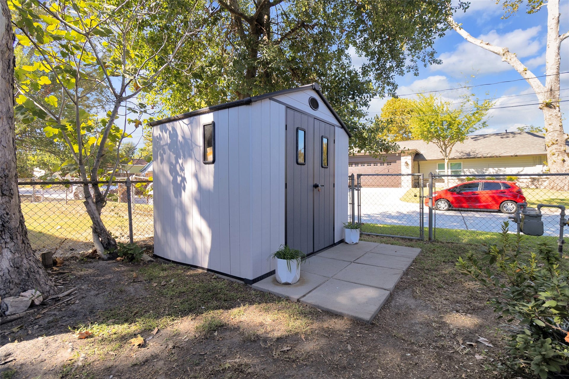 613 N Avenue F, Humble, TX 77338 A charming, well-maintained garden shed sits in a fenced backyard, surrounded by mature trees and enhanced by a paved pathway, offering functional storage space.