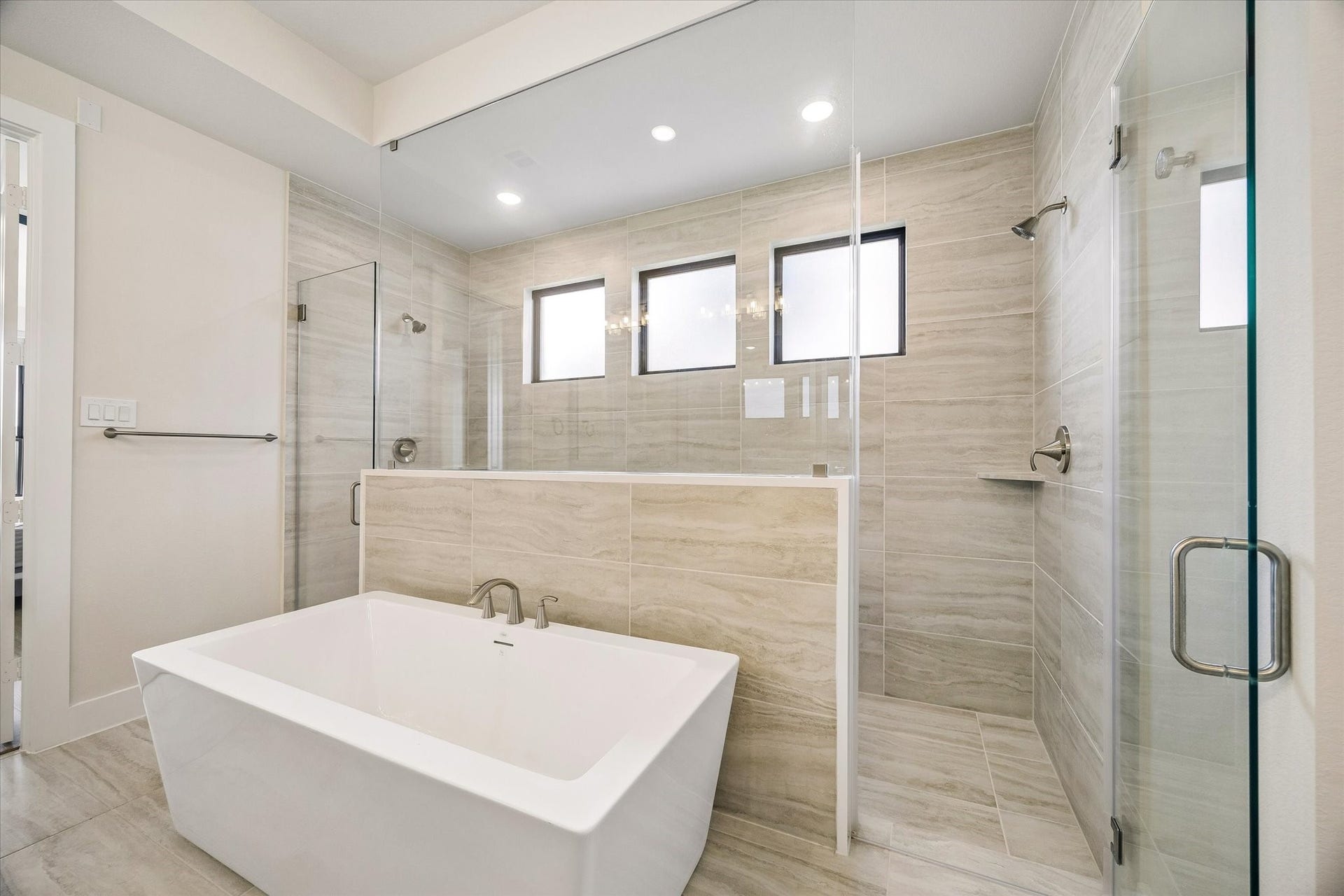 PRIMARY BATH FEATURES A FREESTANDING SOAKING TUB, OVERSIZED WALK-IN SHOWER, FLOOR-TO-CEILING TILE, AND MULTIPLE WINDOWS FOR NATURAL LIGHT.