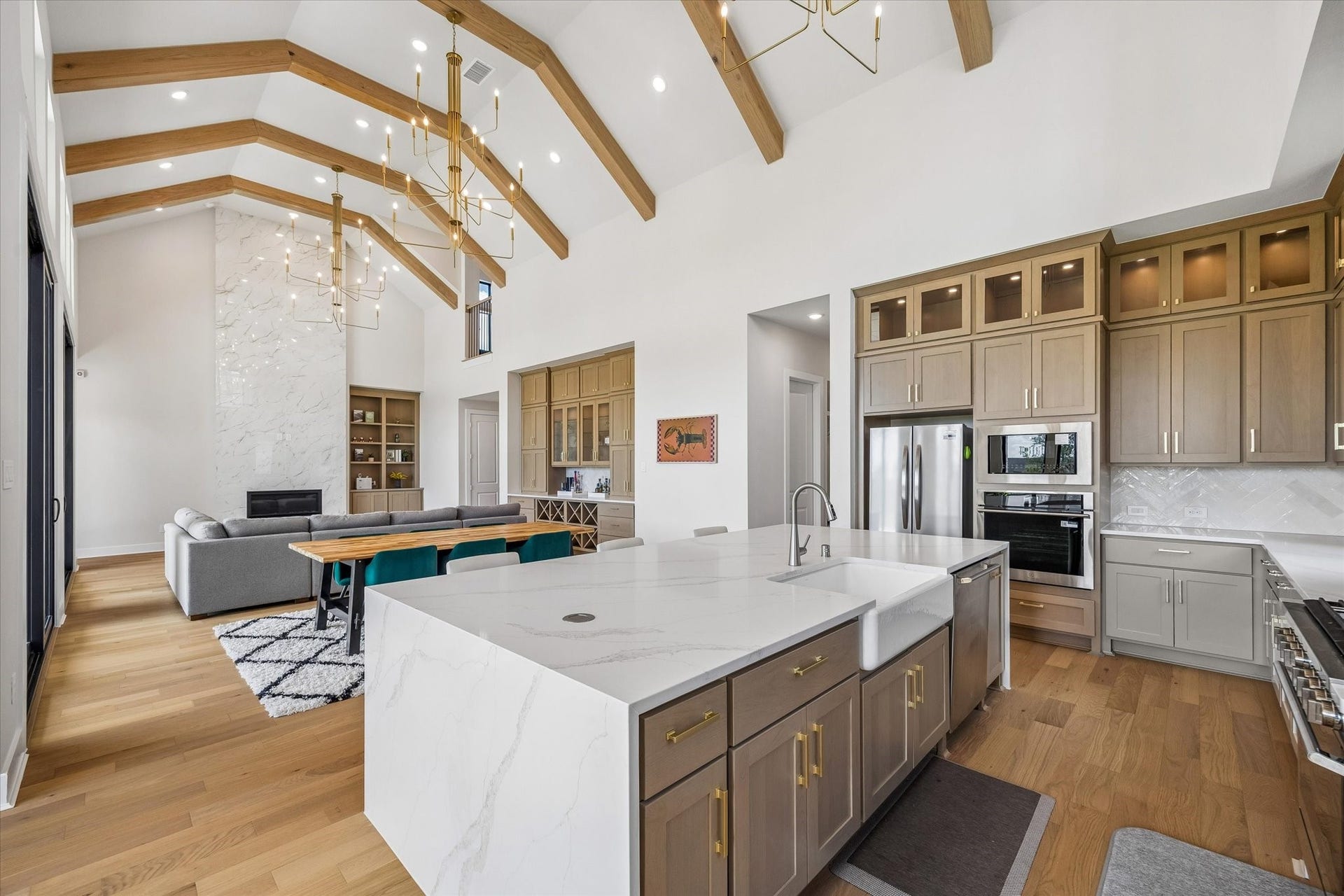 THE OPEN-CONCEPT KITCHEN FEATURES A LARGE WATERFALL ISLAND, FARMHOUSE SINK, QUARTZ COUNTERTOPS, EXTENSIVE CABINETRY, AND STAINLESS STEEL APPLIANCES. VAULTED CEILINGS WITH EXPOSED WOOD BEAMS CONTINUE INTO THE ADJOINING LIVING AREA.