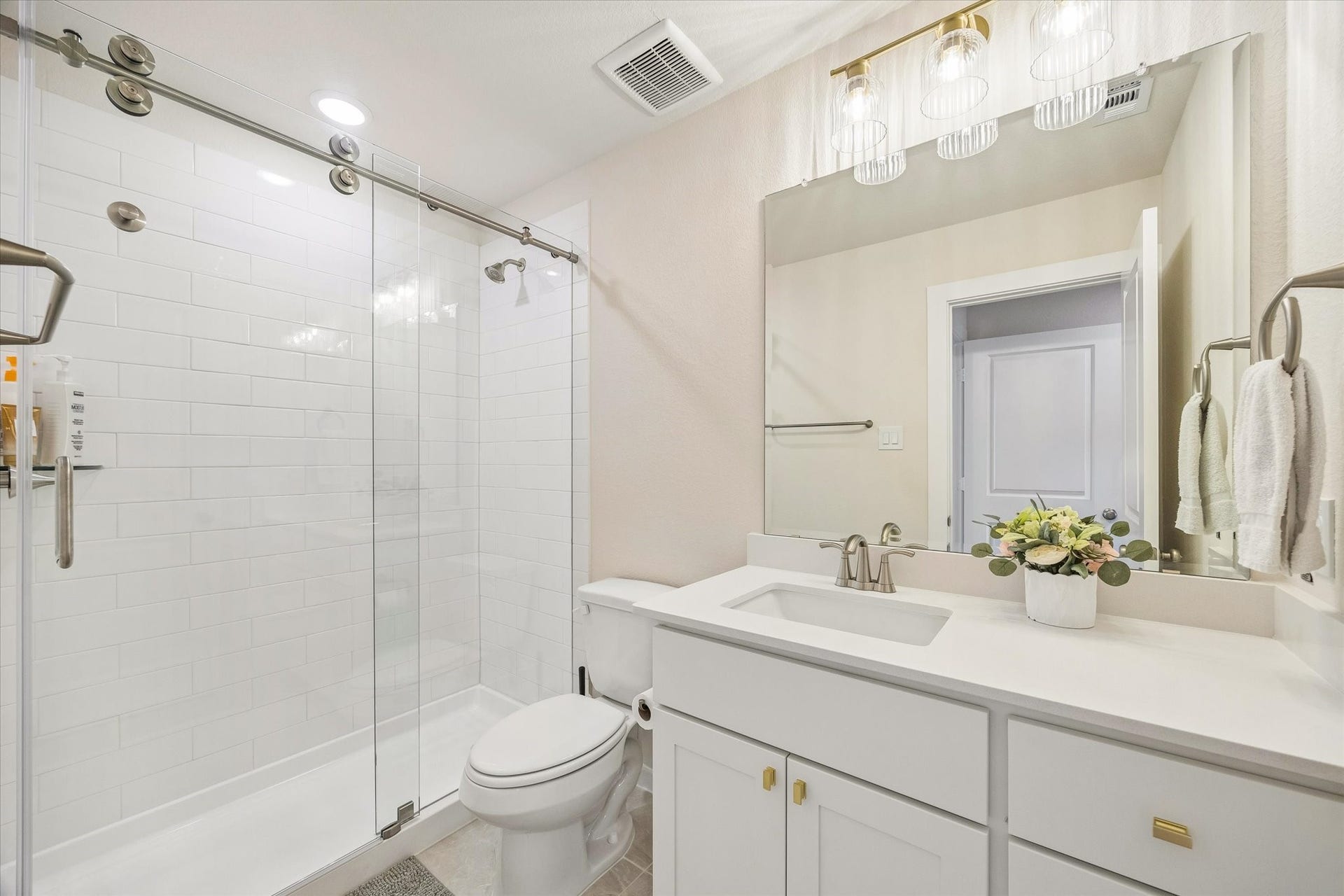 SECONDARY BATH FEATURES A GLASS-ENCLOSED SHOWER, WHITE TILE SURROUND, QUARTZ VANITY, AND MODERN LIGHTING.
