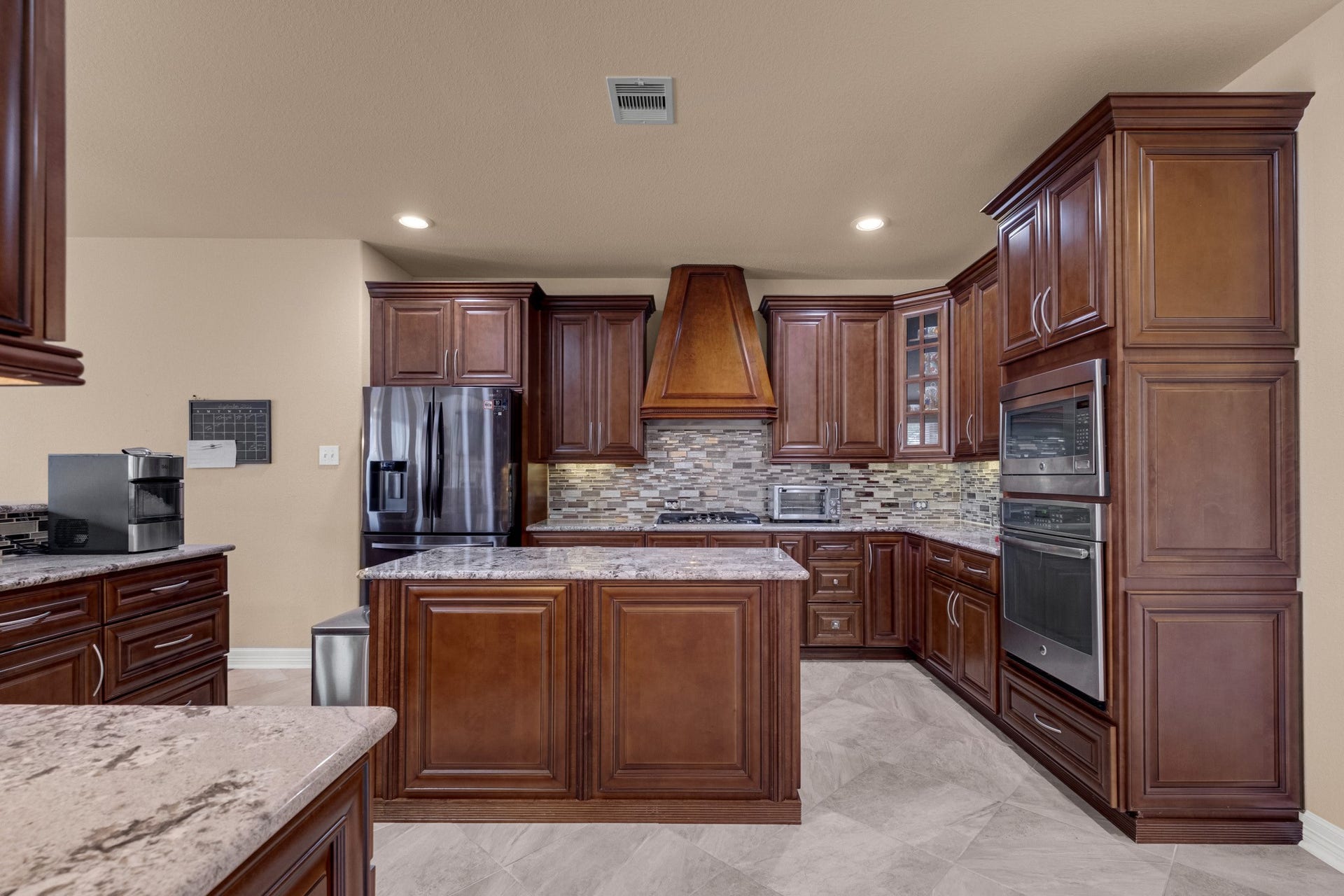 440 Old Orchard Court, Dickinson, TX 77539 The kitchen is dressed in chocolate-glazed cherry cabinetry with a custom tapered wood chimney hood, full-movement granite counters, a linear glass and travertine mosaic backsplash, and a stainless wall oven with microwave above, set into a full-height cabinet tower.