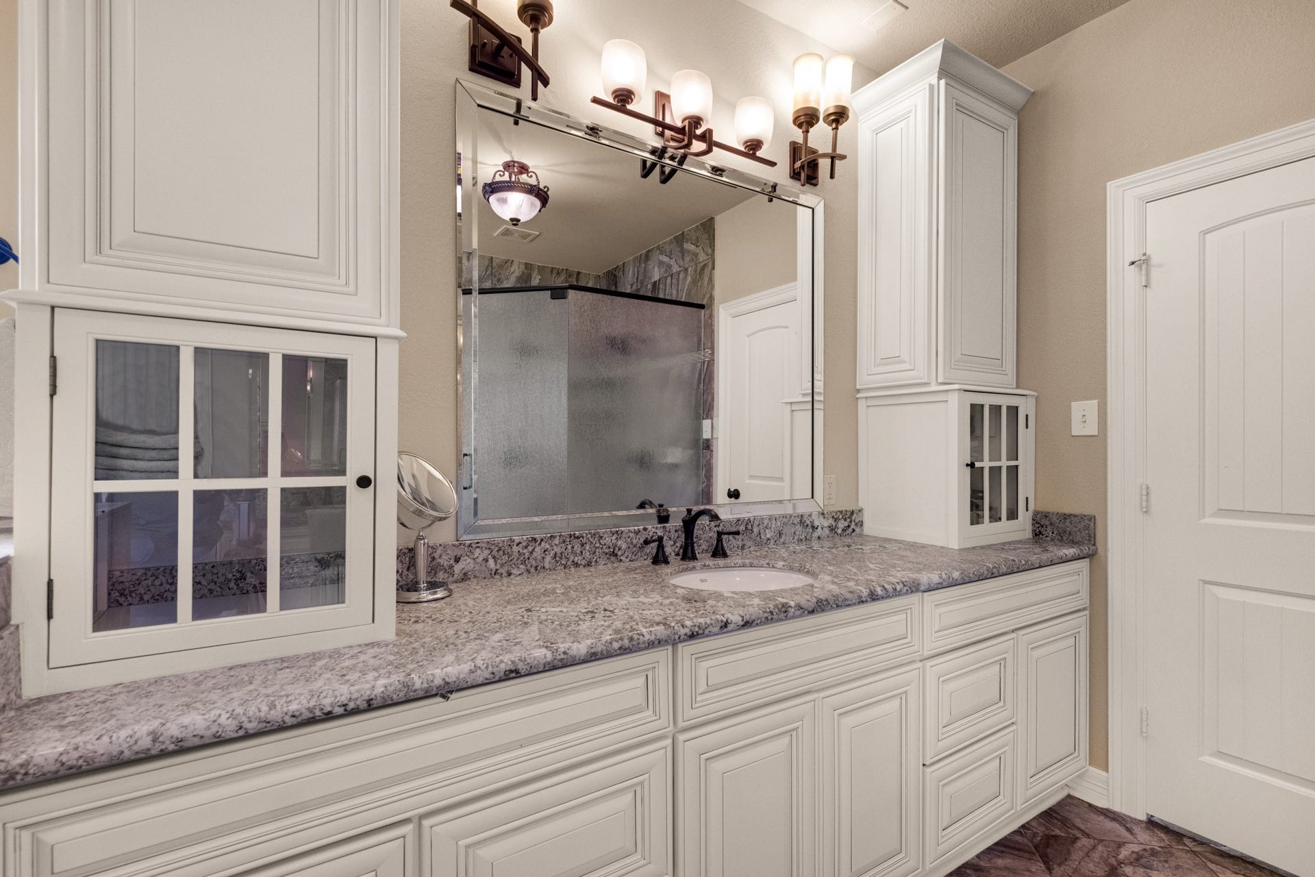 440 Old Orchard Court, Dickinson, TX 77539 The vanity wall stretches long in granite with framed mirror, sconce lighting, and glass-front cabinetry for display and storage together.