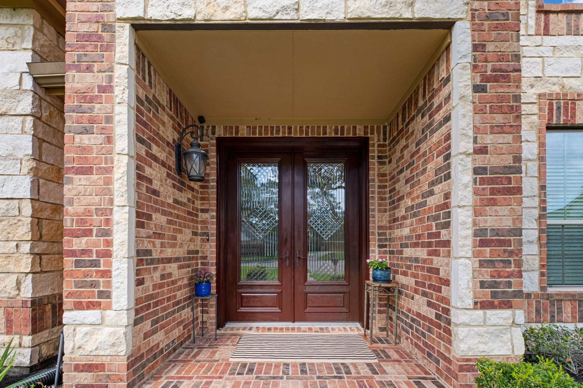 440 Old Orchard Court, Dickinson, TX 77539 The covered entry frames a mahogany-toned double door set with wrought-iron diamond-lattice glass, standing between cast stone quoins and running-bond brick, with a herringbone brick porch underfoot.