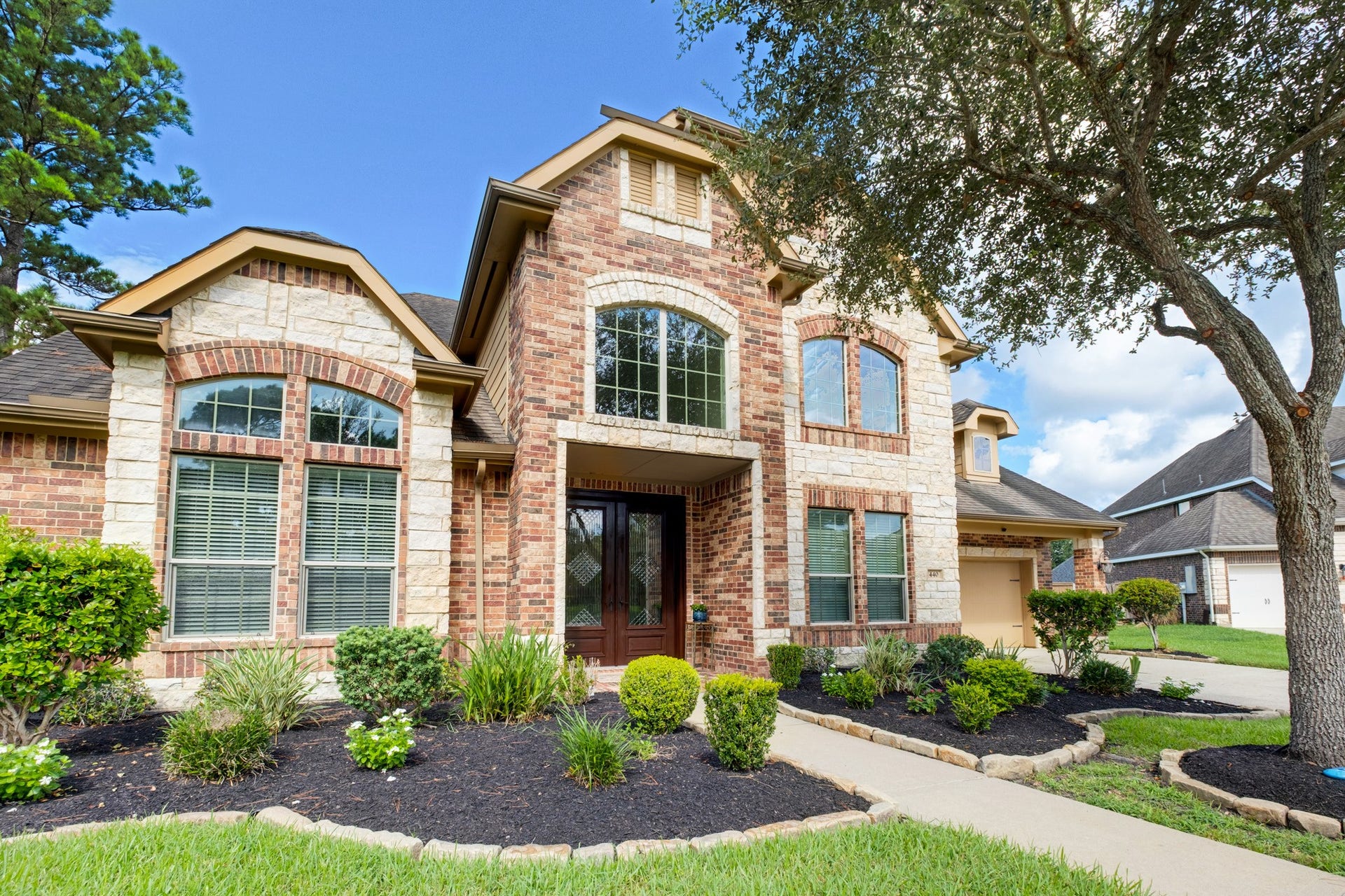 440 Old Orchard Court, Dickinson, TX 77539 By daylight the craftsmanship reads even clearer: arched windows crowned with soldier-course brick and cast stone surrounds, a stained double entry door set with wrought-iron diamond-lattice glass, and a curved walkway winding through layered beds to the front step.