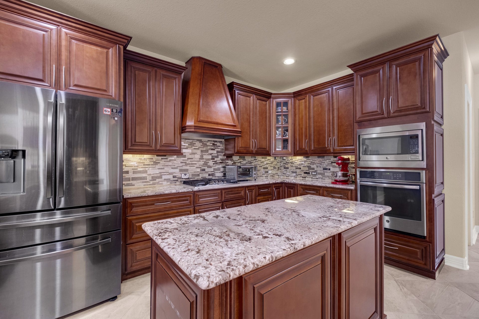 440 Old Orchard Court, Dickinson, TX 77539 The custom tapered wood chimney hood crowns a gas cooktop set in full-movement granite, with a stainless microwave and wall oven stacked in the cabinet tower and a French-door refrigerator alongside.