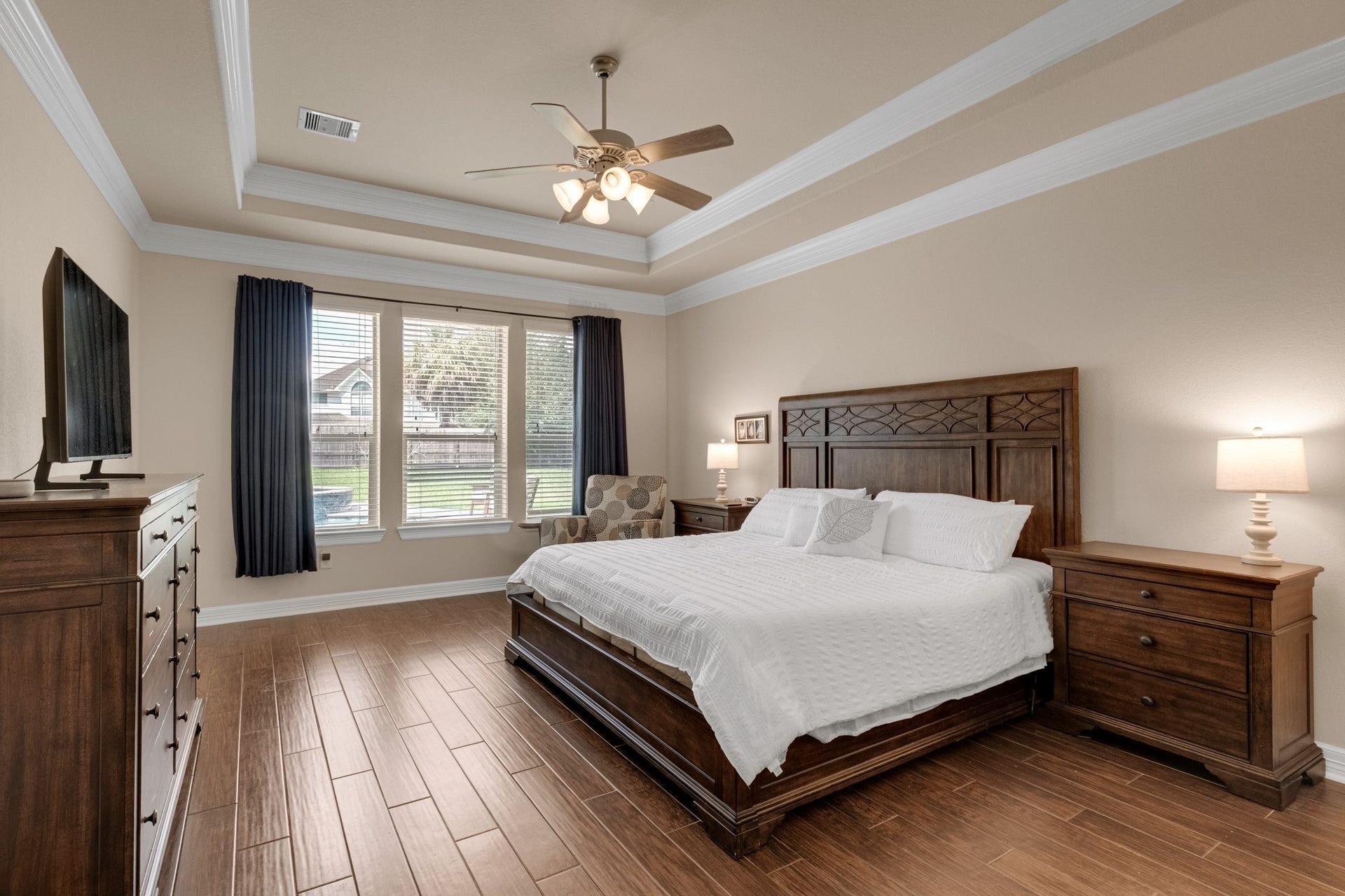 440 Old Orchard Court, Dickinson, TX 77539 The primary suite retreats behind a stepped tray ceiling trimmed in crown molding, with mahogany wood tile underfoot and a wide window bank framing the shaded backyard.
