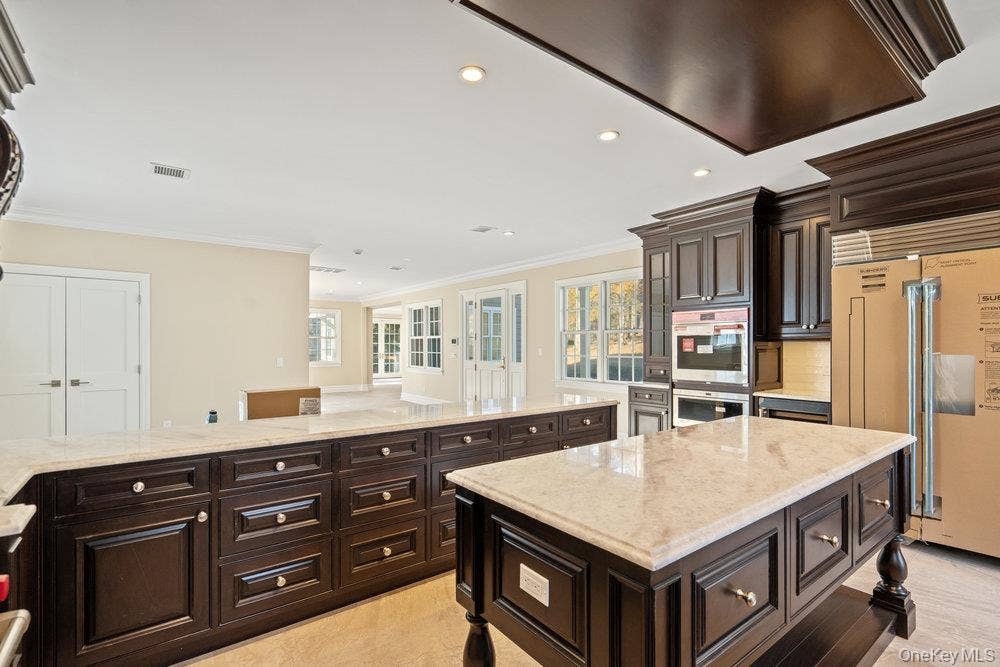 89 Mill River Road, Upper Brookville, NY 11771 Kitchen with dark brown cabinetry, exhaust hood, paneled built in fridge, light stone countertops, and stainless steel double oven