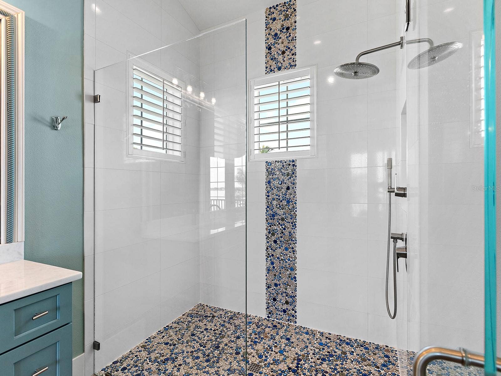 4024 11TH STREET COURT W, Palmetto, FL 34221 Luxe walk-in shower oasis with a vibrant blue and tan river rock mosaic feature wall, matching pebbled floor, a wall-mounted showerhead, and privacy-enhancing plantation shutters