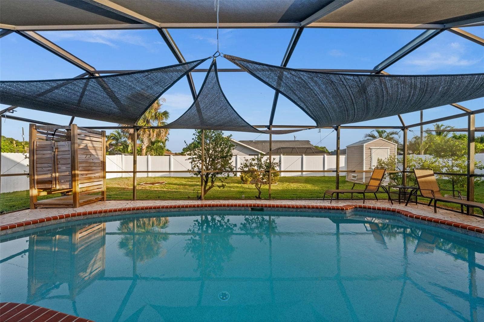 Screen-enclosed pool area featuring a brick-tiled perimeter, overhead fabric shade sails, and a freestanding wood-plank outdoor shower enclosure. Notice the storage shed and the custom shower?